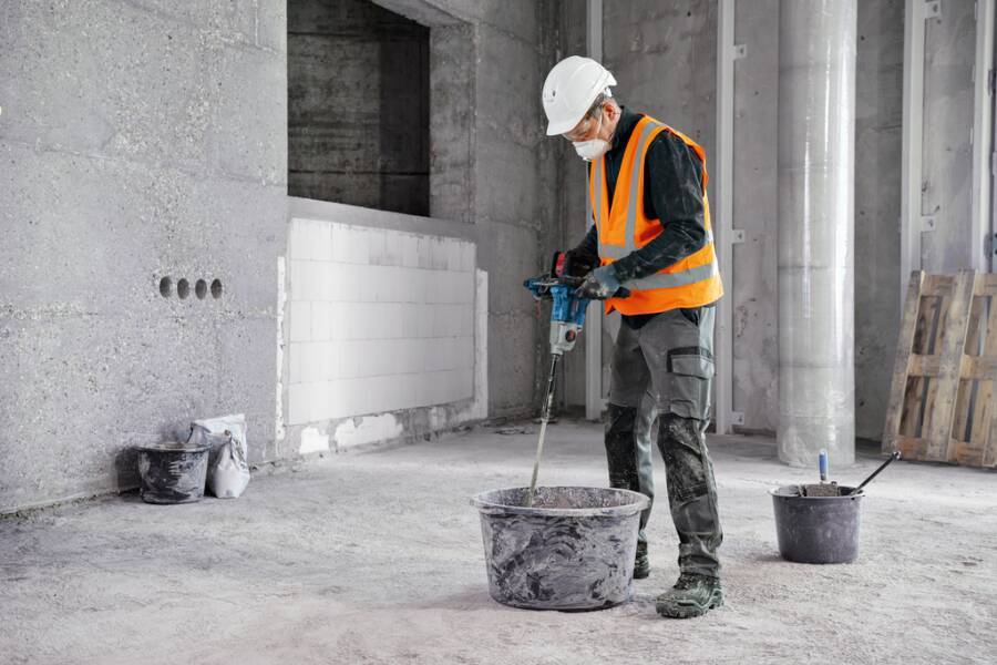 Ein Bauarbeiter mit Schutzhelm, Warnweste und Maske benutzt einen Bohrer, um Materialien in einem Eimer auf einer Baustelle zu mischen.