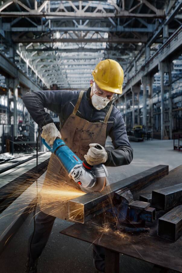 Arbeiter mit Sicherheitsausrüstung schleift Metall in einer großen Industriehalle. Funken sprühen vom Winkelschleifer und unterstreichen die intensive manuelle Arbeit.