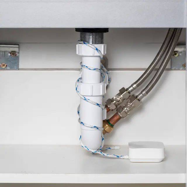 Unterschrank-Rohrleitungen mit einem weißen Abflussrohr, das mit blau-weißem Seil umwickelt ist, und zwei geflochtenen Versorgungsschläuchen, die auf der rechten Seite angeschlossen sind, neben einem kleinen weißen Sensorgerät.