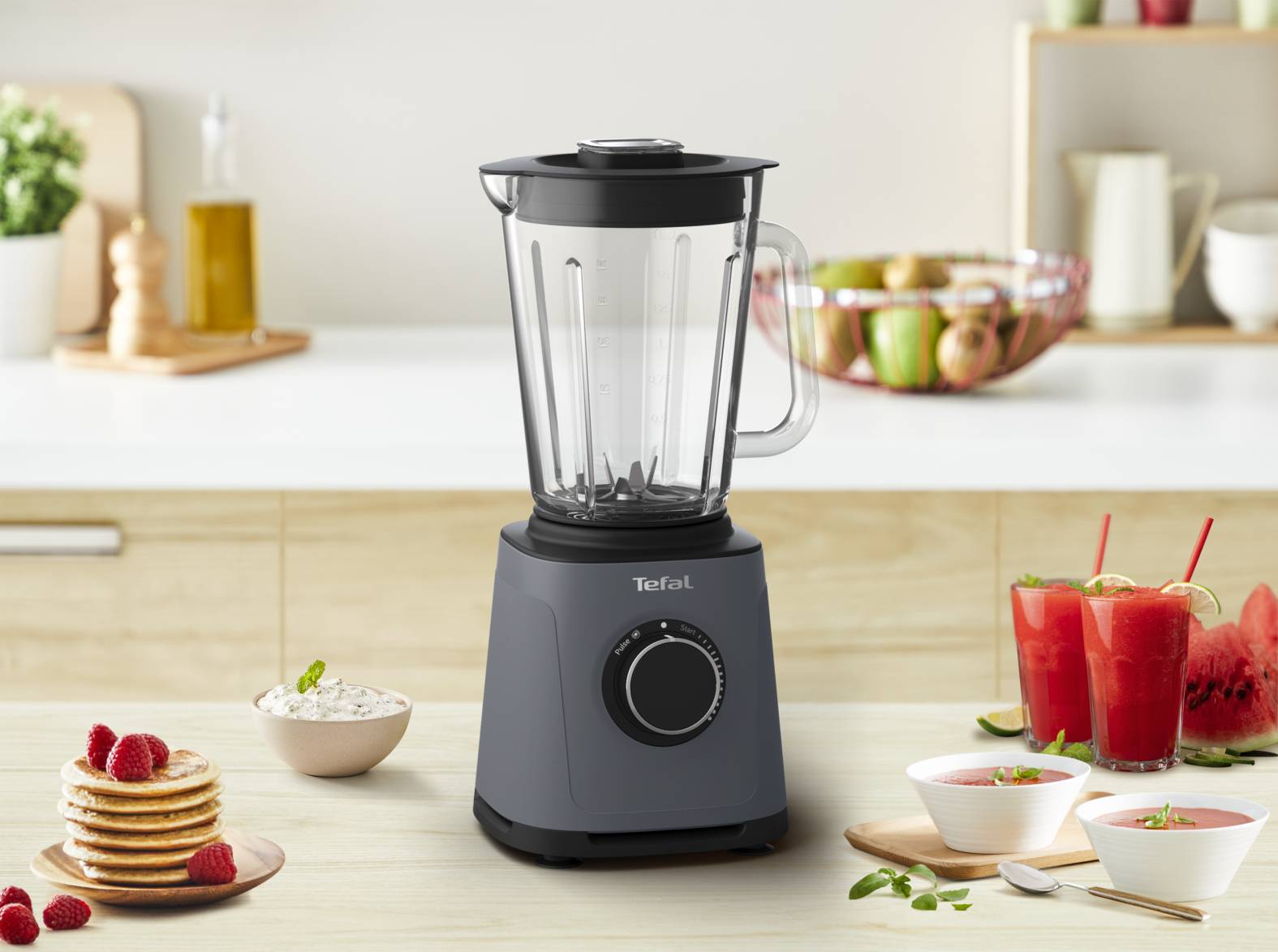 Ein Standmixer sitzt zentriert auf einer Küchenarbeitsplatte, mit Schüsseln voller Obst und Smoothies drumherum, was auf Essenszubereitung oder die Herstellung von Getränken hindeutet.