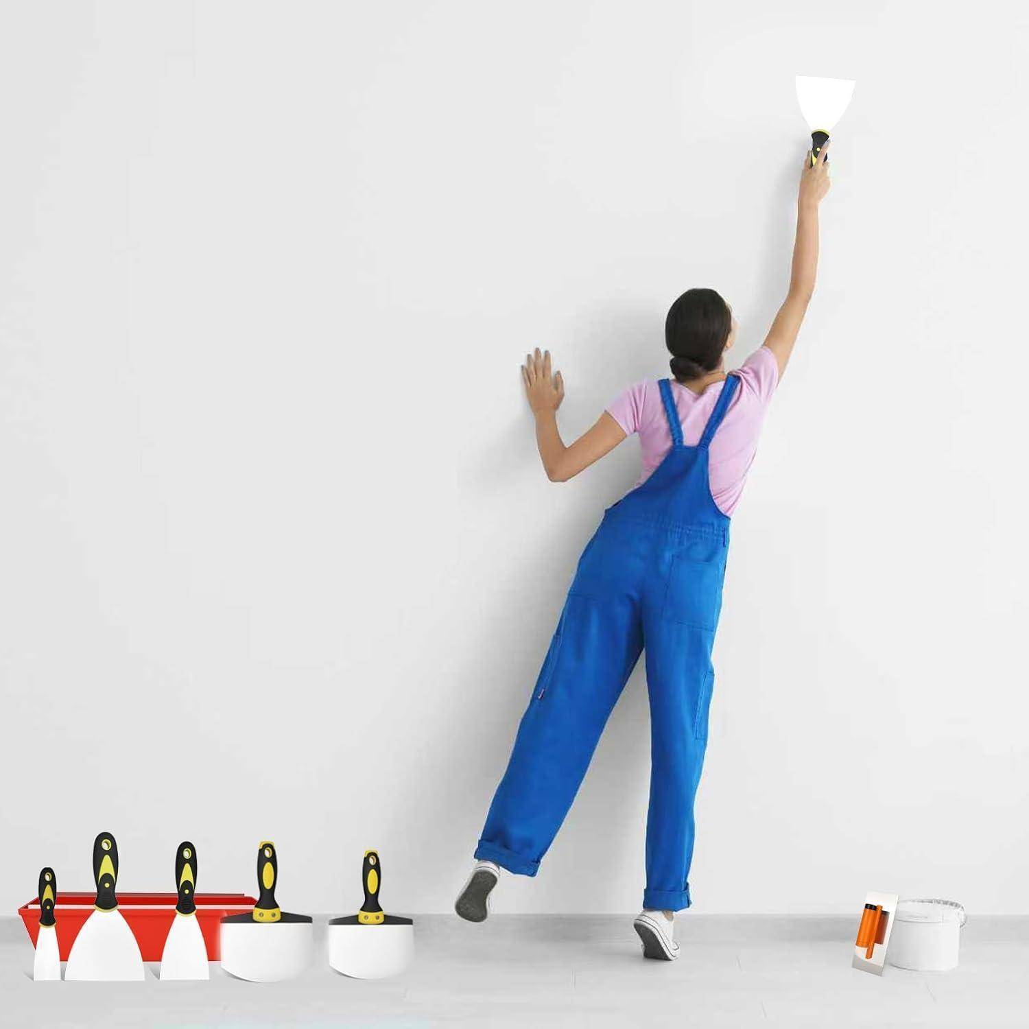 Eine Person in blauen Overalls trägt mit einer Spachtel Spachtelmasse auf eine weiße Wand auf. Verschiedene Werkzeuge und Behälter befinden sich in der Nähe auf dem Boden.