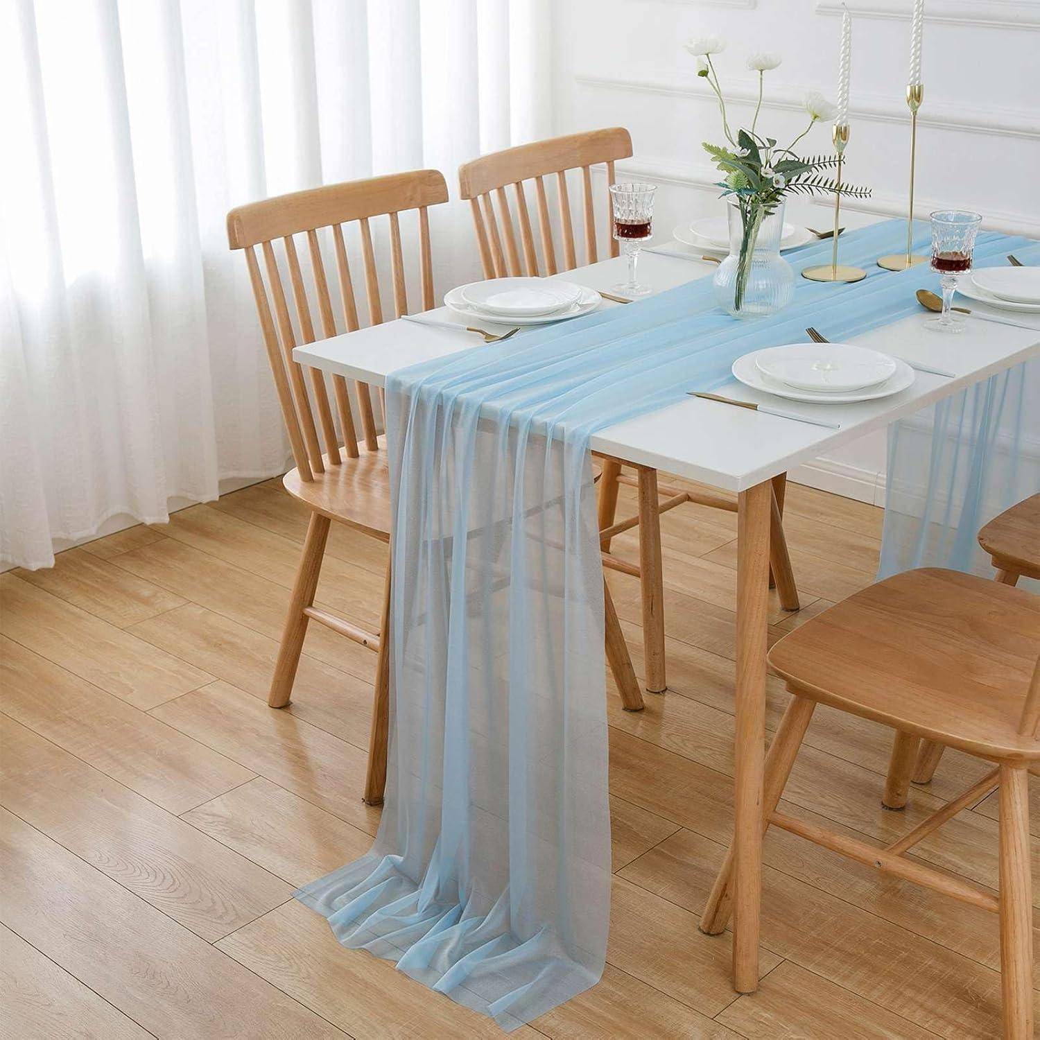 Ein Esstisch, bestuhlt mit Holzstühlen, einem hellblauen Tischläufer, weißen Tellern und Gläsern. Eine Vase mit weißen Blumen steht auf dem Tisch.