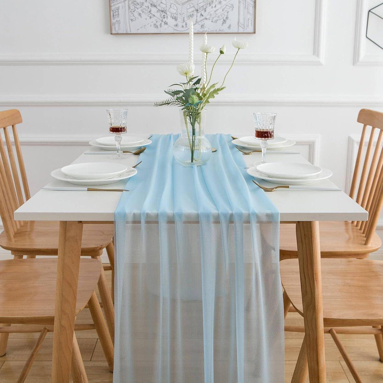Ein Esstisch, gedeckt für vier Personen, mit hellen Holzstühlen, weißen Tellern und Gläsern mit roter Flüssigkeit. Ein hellblauer Läufer und ein florales Mittelstück verleihen dem Arrangement Eleganz.