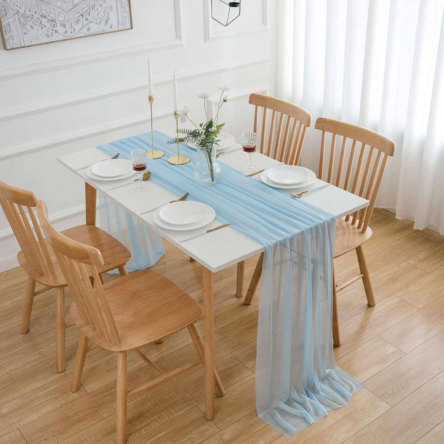 Ein Holz-Esstisch für vier Personen mit einem hellblauen Tischläufer, weißen Tellern und Blumen in Vasen. Natürliches Licht vom Fenster.