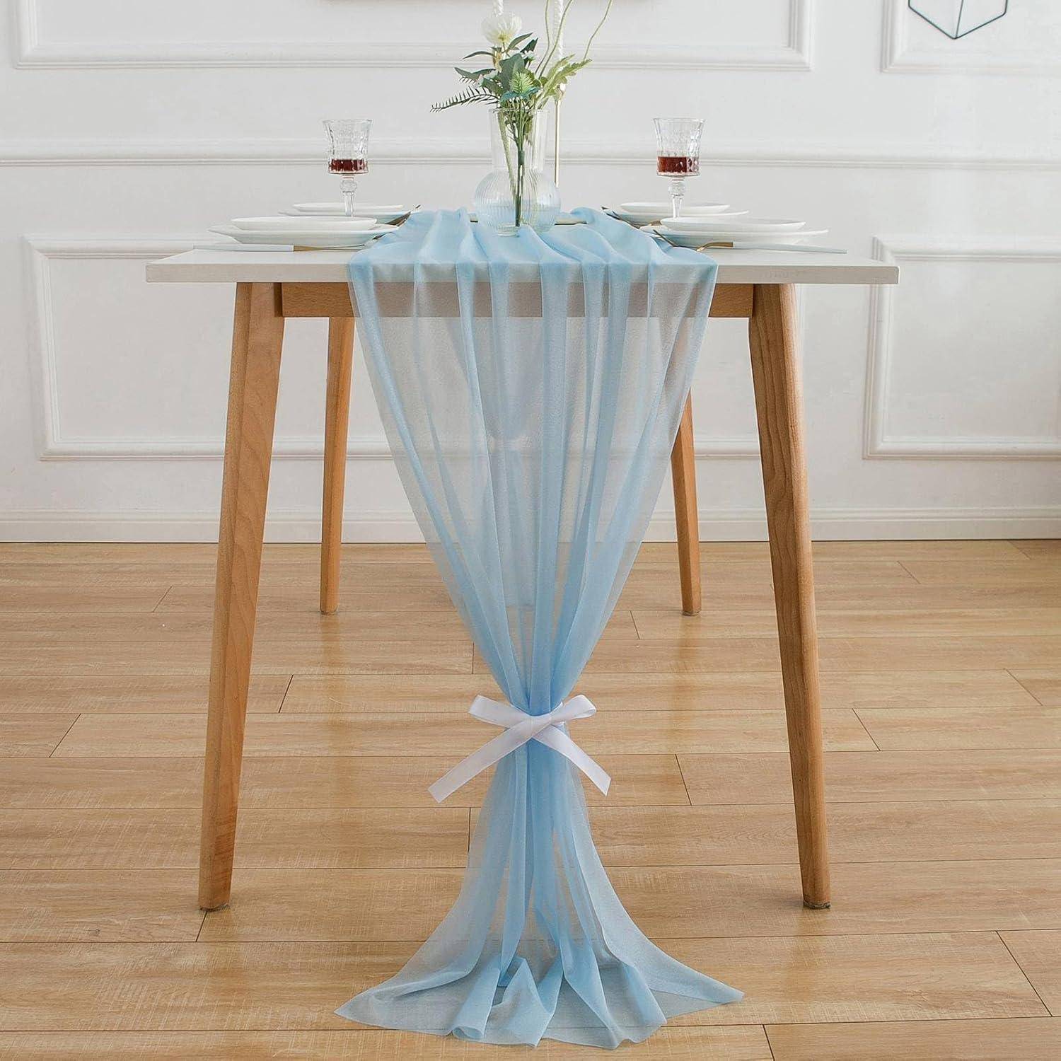 Ein quadratischer Tisch mit Holzbeinen ist elegant geschmückt mit einem hellblauen durchsichtigen Tischläufer, der mit einem weißen Band gebunden ist, und darauf platziert eine Blumenvase sowie Gläser mit Rotwein.