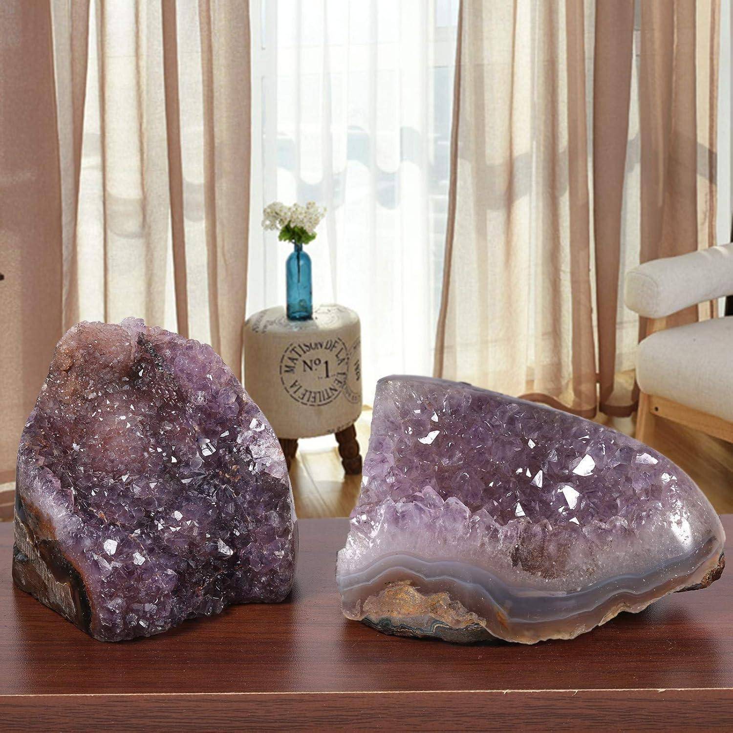 Zwei große Amethystkristalle mit tiefen Purpurtönen sitzen auf einem Holztisch in einem Raum mit beigen Vorhängen und einer kleinen Vase auf einem runden Beistelltisch.