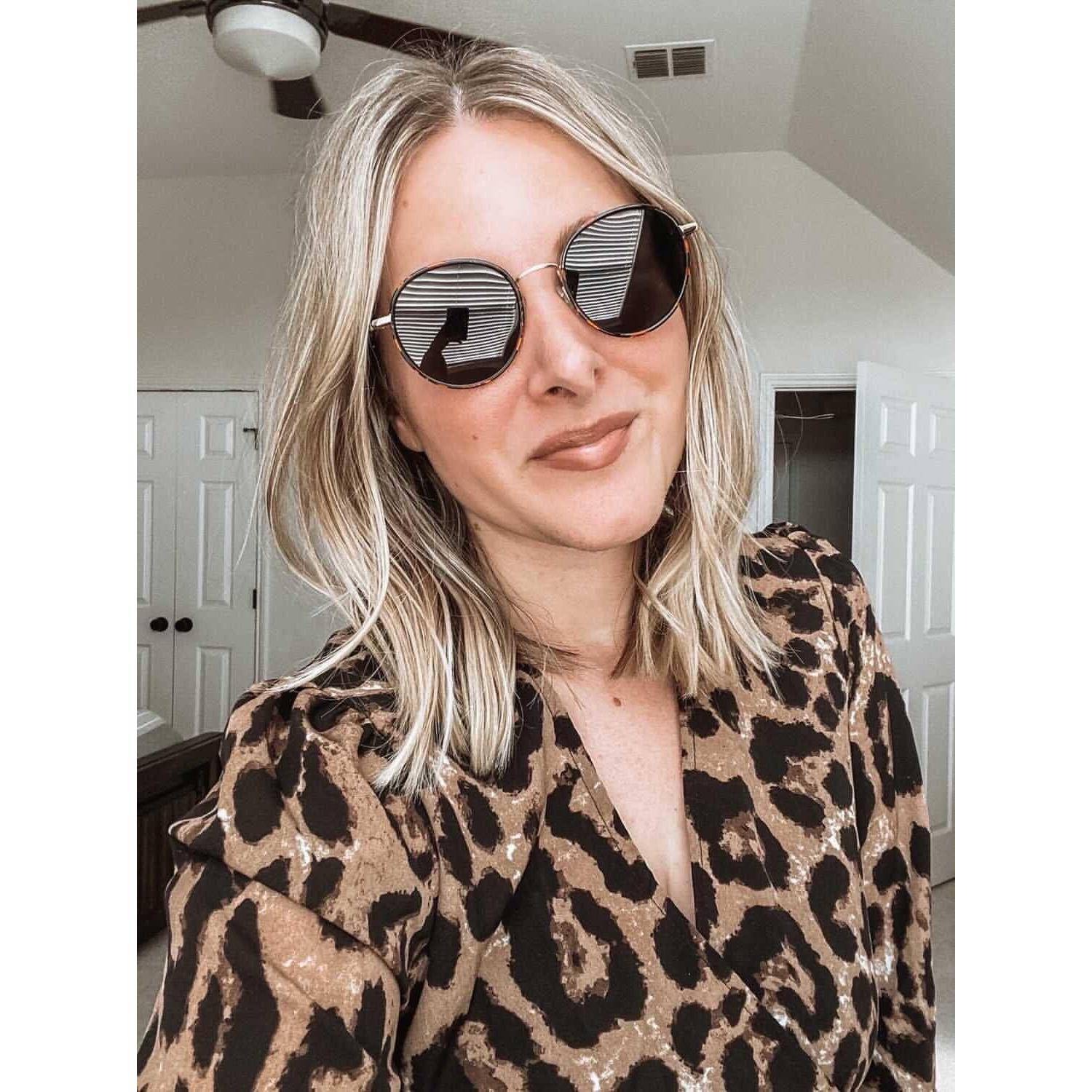 Eine Person mit blonden Haaren und großen runden Sonnenbrille lächelt in einem Raum in eine Kamera, trägt eine gemusterte Bluse.