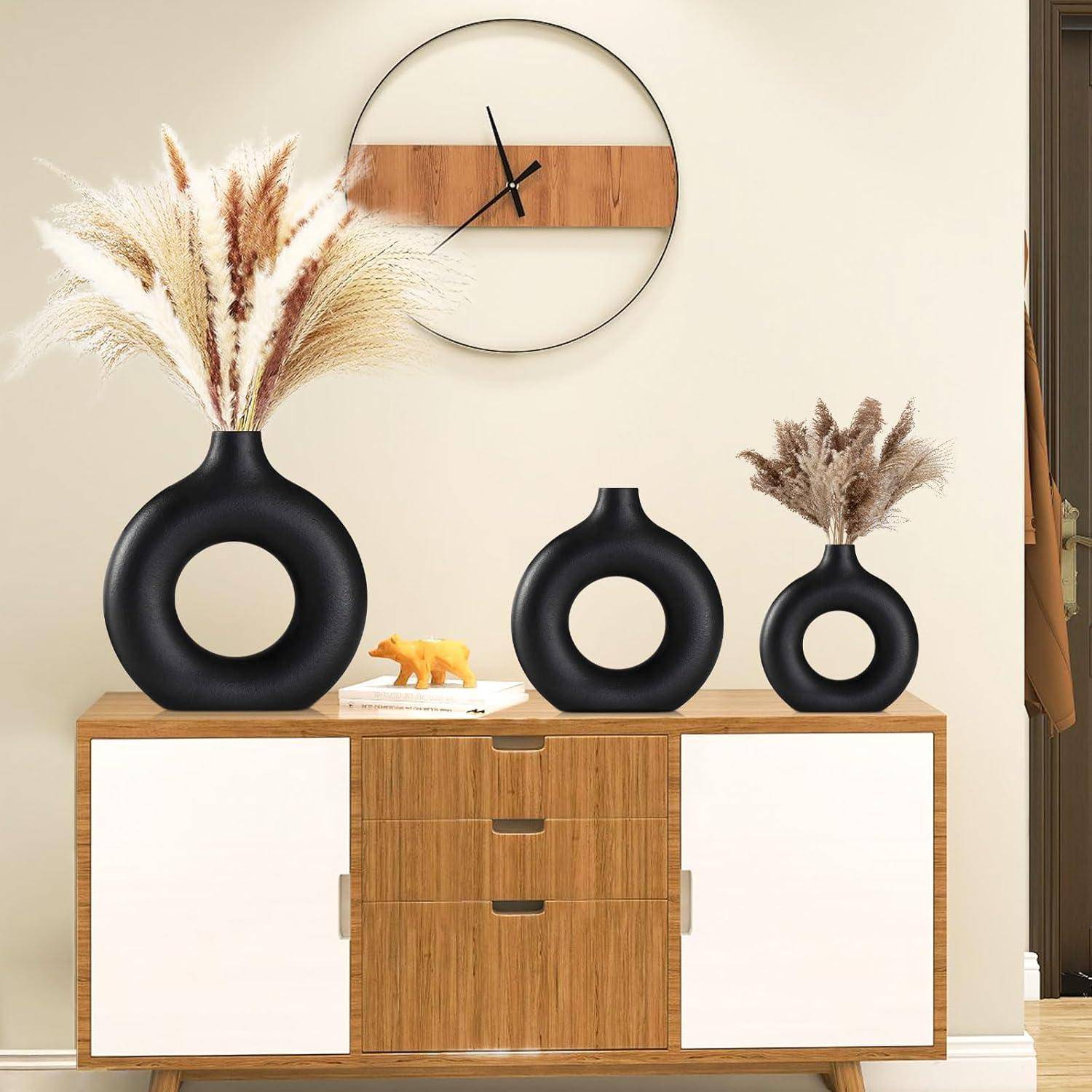 Ein modernes Wohnzimmer mit einem Holzsideboard, drei runden schwarzen Vasen mit Pampasgras und einer dekorativen minimalistischen Uhr an der Wand.