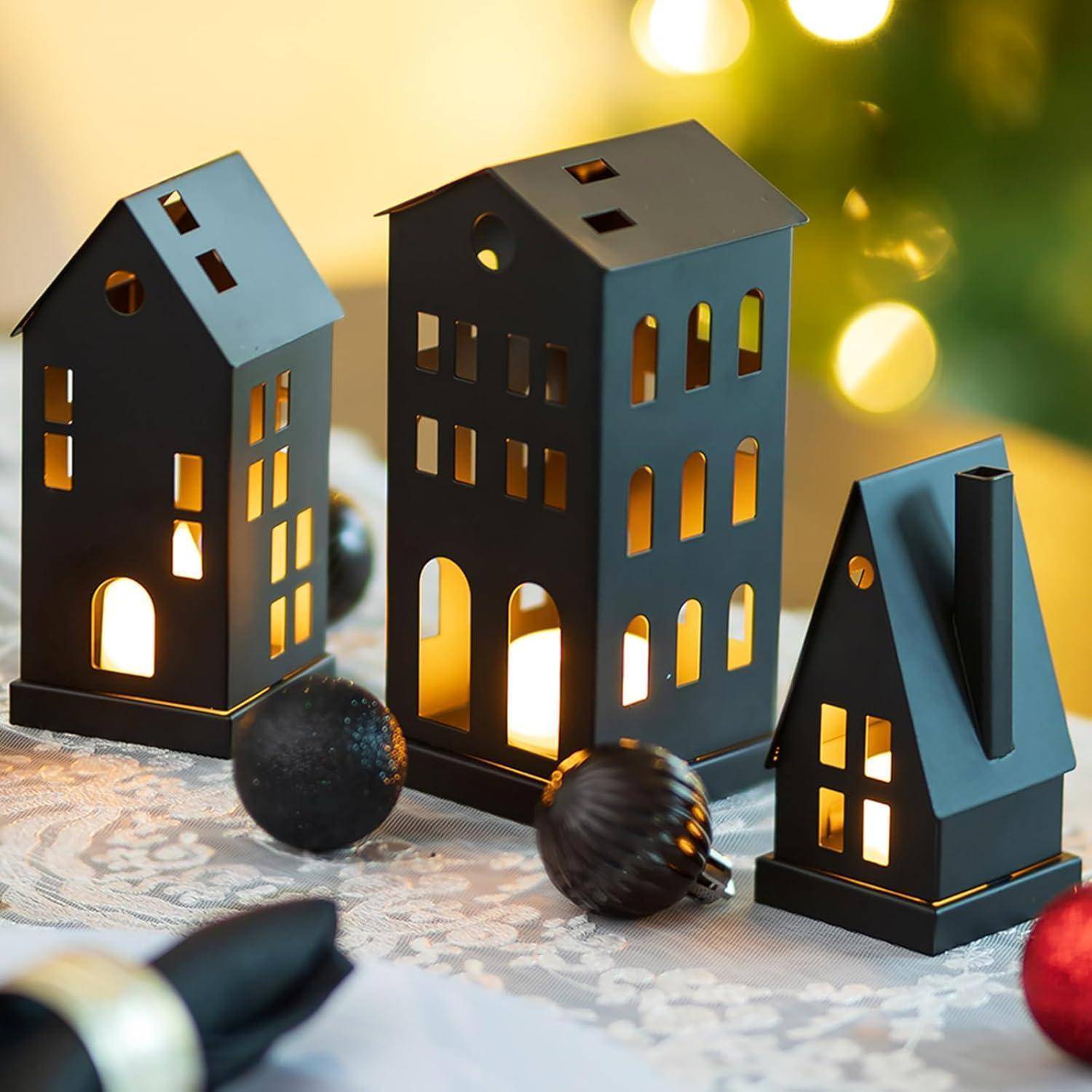 Miniaturhauslampen mit brennenden Kerzen im Inneren, auf einem Spitzentischtuch platziert, neben dunklen Ornamenten vor einem unscharfen festlichen Hintergrund.