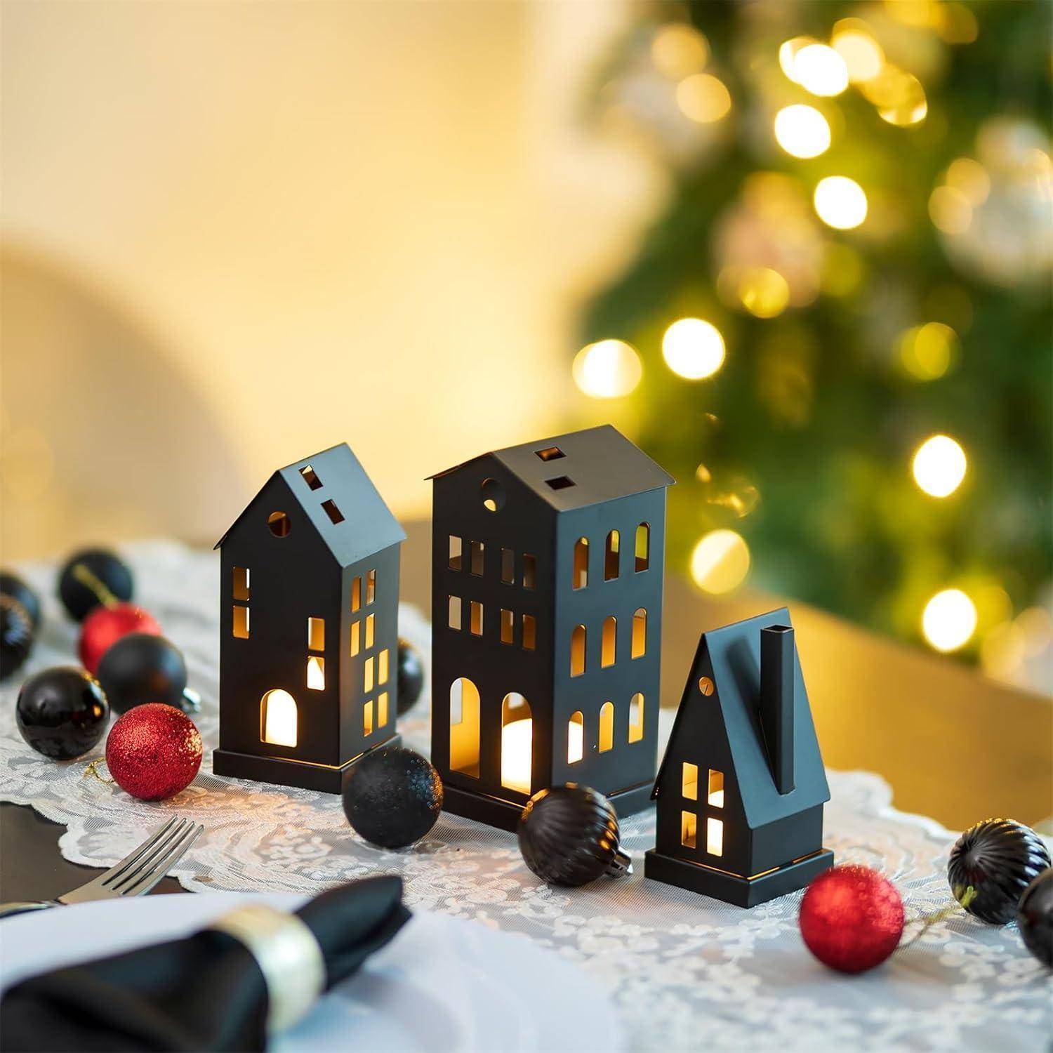 Miniaturhauslaternen mit brennenden Kerzen, umgeben von roten und schwarzen Ornamenten auf einem Tisch; verschwommene Weihnachtsbaumlichter im Hintergrund.