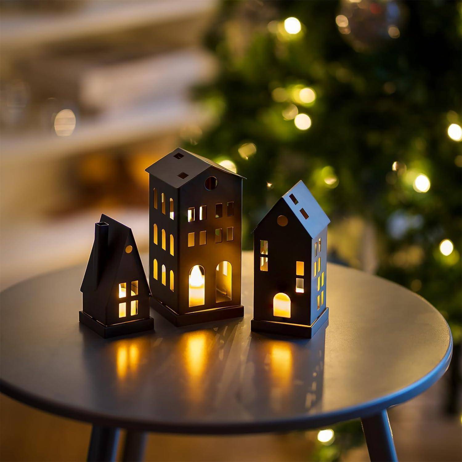 Eiserne Kerzenhalter in Form von Häusern, die von Kerzen von innen beleuchtet werden, stehen auf einem grauen Tisch mit einem unscharfen Weihnachtsbaum und Lichtern im Hintergrund.