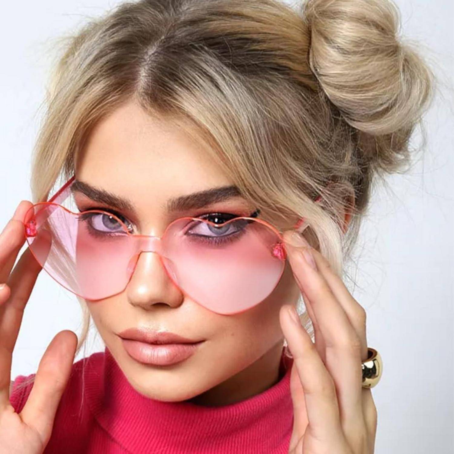 Eine Person mit blonden Haaren in zwei Haarknoten trägt herzförmige Sonnenbrille mit rosa Tönung und ein rosa Oberteil und blickt direkt in die Kamera.