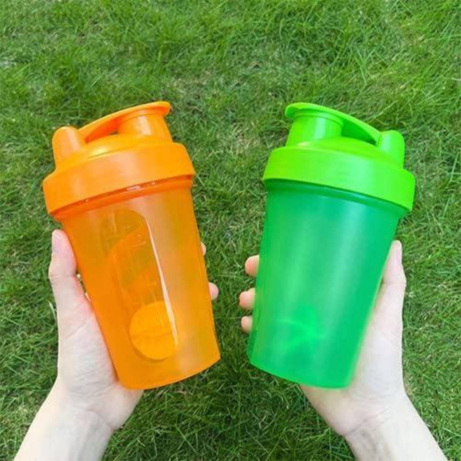 Zwei Hände halten eine leuchtend orange Shakerflasche links und eine leuchtend grüne Shakerflasche rechts, vor einem grasigen Hintergrund.