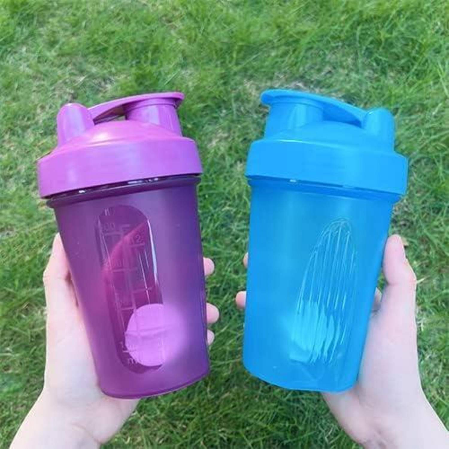 Zwei Hände, die einen lila Shaker auf der linken Seite und einen blauen Shaker auf der rechten Seite vor einem Rasenhintergrund hochhalten.