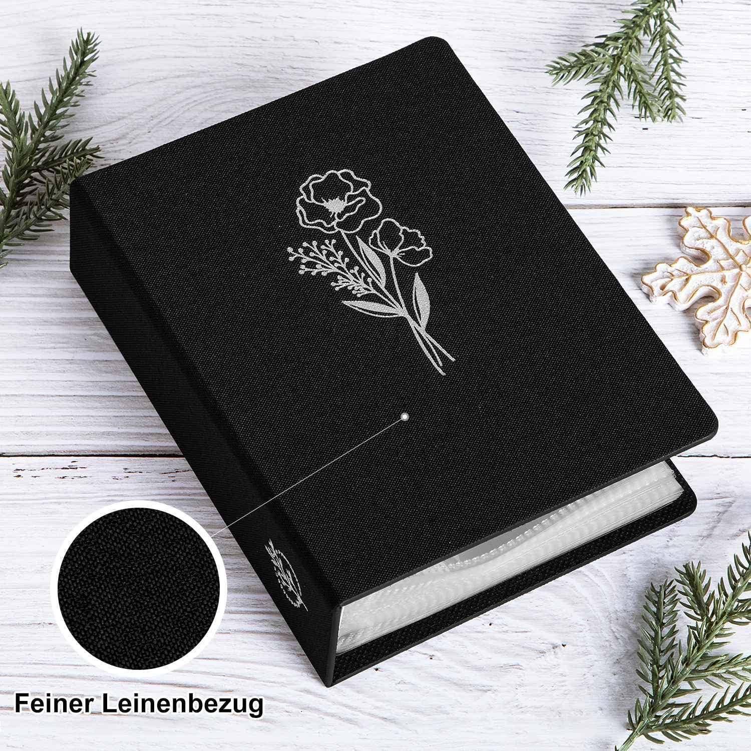 Ein schwarzes Buch mit einem silbernen Blumendesign auf dem Einband, umgeben von Tannenzweigen auf einer weißen Holzoberfläche. Deutscher Text lautet 