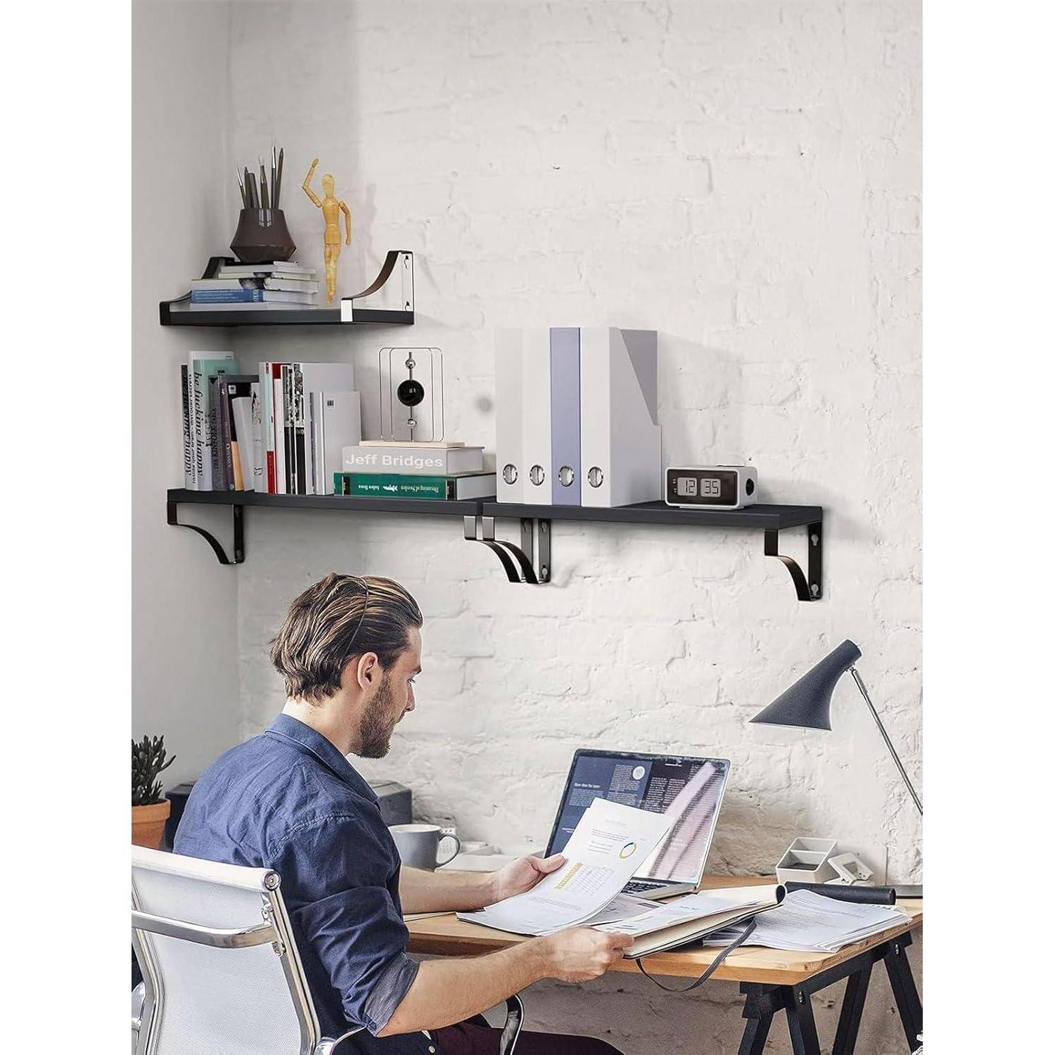 Eine Person sitzt an einem Schreibtisch und arbeitet an einem Laptop, während sie ein Dokument liest. An einer weißen Ziegelwand sind Regale mit Büchern und Ordnern angebracht.