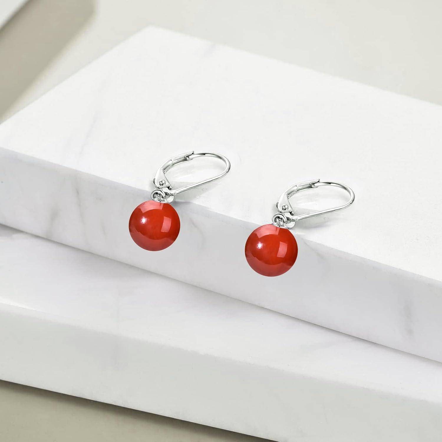 Ein Paar silberne Creolen mit roten kugelförmigen Perlen ruht auf einer weißen Marmoroberfläche und präsentiert ihr elegantes, minimalistisches Design.