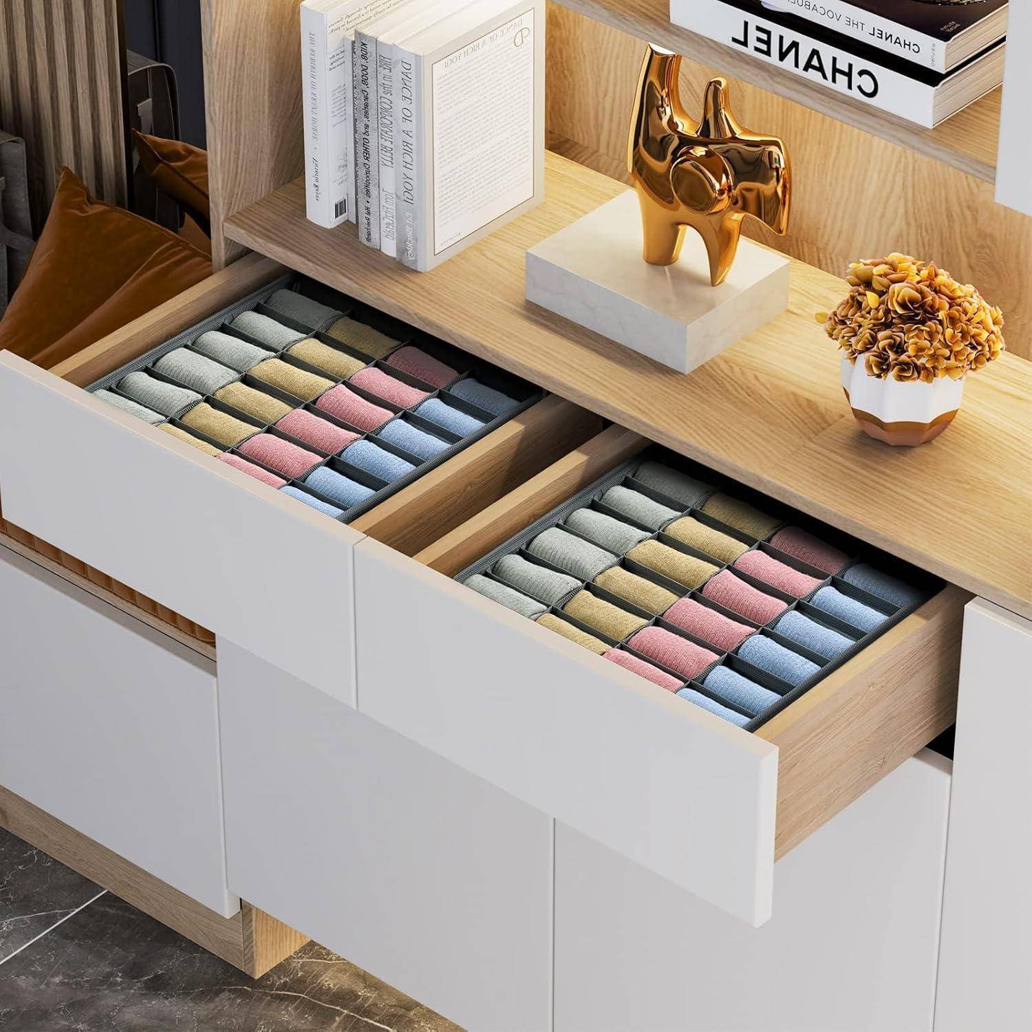 Holzregal mit Büchern, Dekorationselementen und zwei offenen Schubladen, die mit sorgfältig angeordneten, farblich aufeinander abgestimmten aufgerollten Kleidungsstücken gefüllt sind.