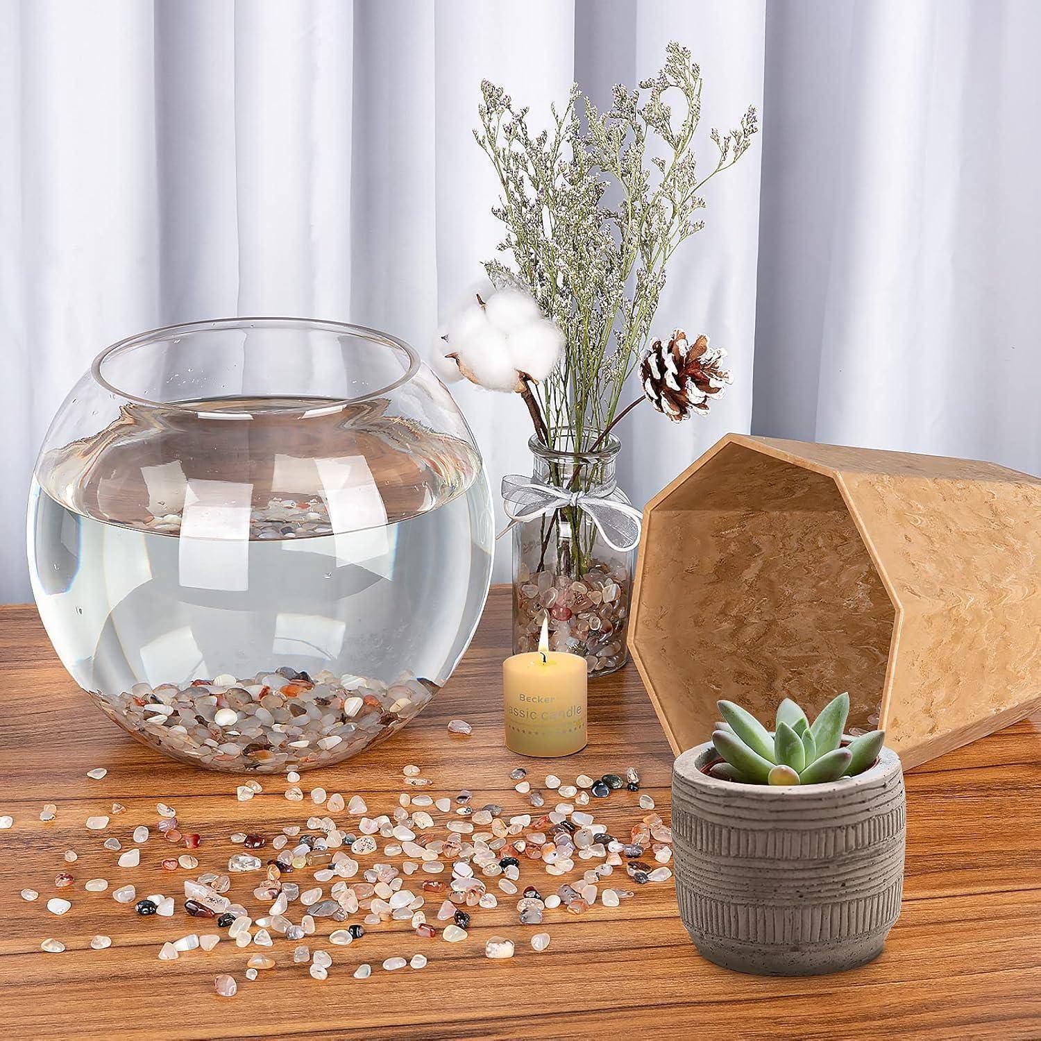 Eine Glasschüssel, gefüllt mit Wasser und Kieseln, steht auf einem Holztisch, umgeben von getrockneten Blumen, einer Kerze und einer Sukkulente im Topf.