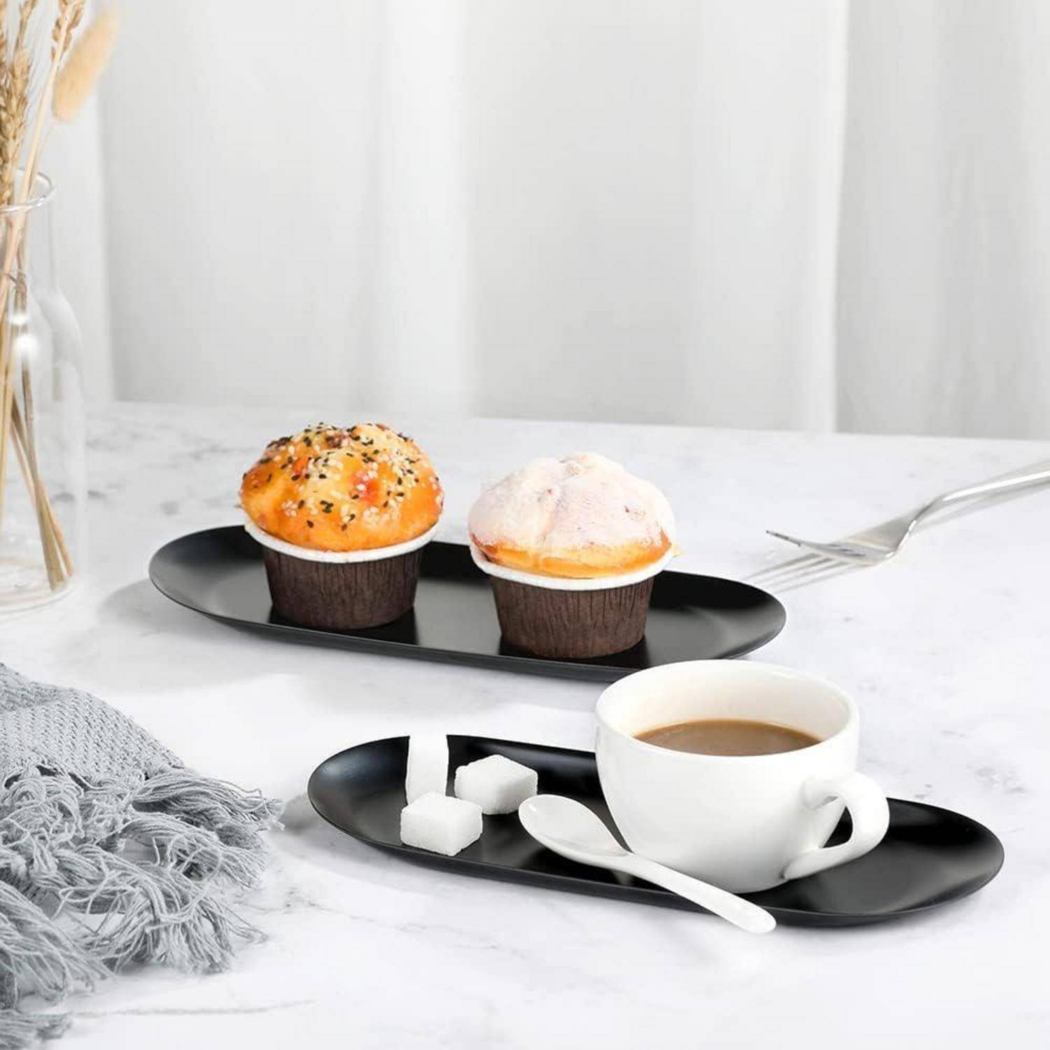 Zwei Muffins auf einem schwarzen Teller neben einer Tasse Kaffee mit Würfelzucker auf einem schwarzen Tablett auf einem Marmortisch.