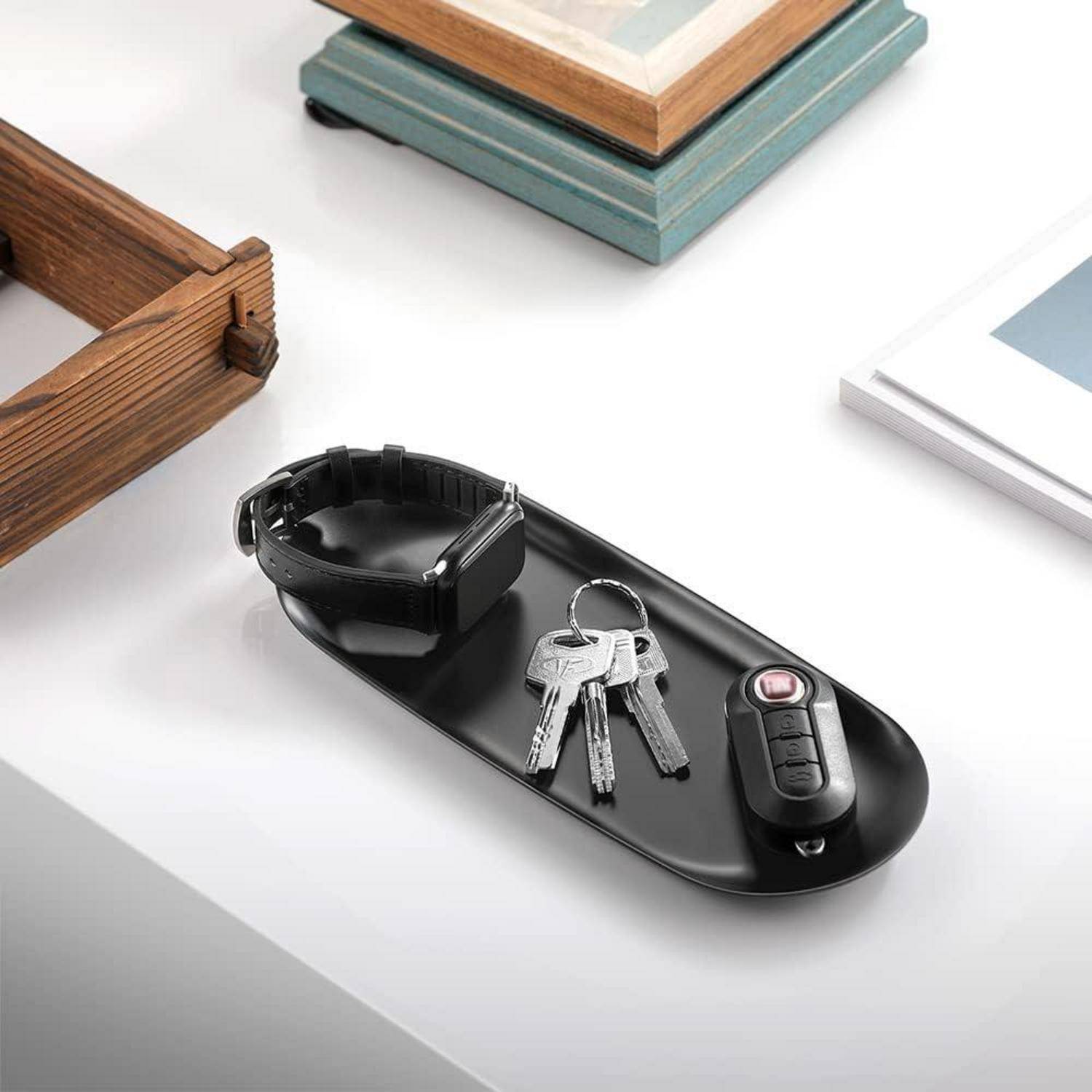 Ein schwarzes Tablett trägt eine Smartwatch, einen Schlüsselbund und eine Autofernbedienung auf einem weißen Schreibtisch neben hölzernen und blauen Büchern.