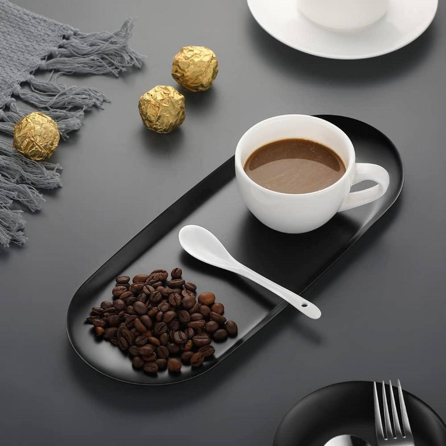 Eine Tasse Kaffee auf einem schwarzen Tablett mit einem Löffel, Kaffeebohnen und Schokoladen. Ein graues Tuch und ein weißer Teller mit einer Tasse im Hintergrund.