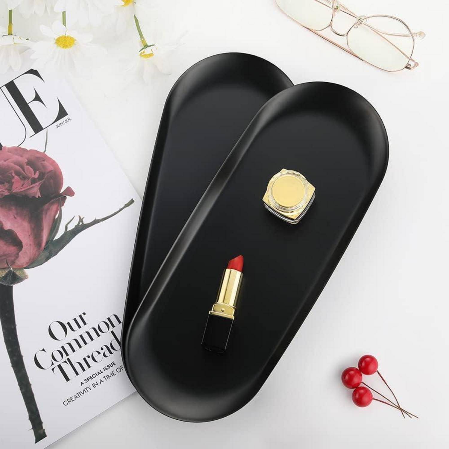 Lippenstift und ein kleiner Behälter auf schwarzen Tabletts; begleitet von einer Modezeitschrift, Brillen und kleinen dekorativen Beeren.