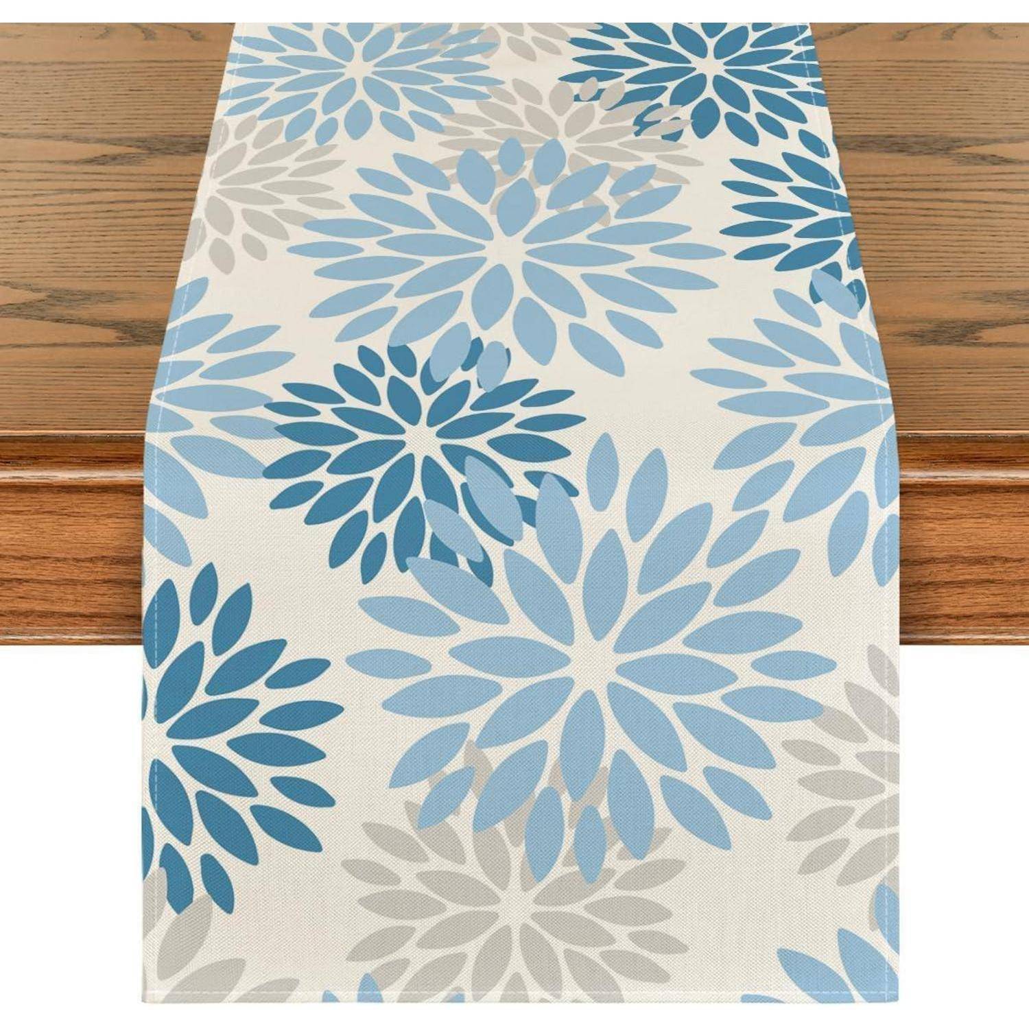 Ein Tischläufer mit einem Blumenmuster, das große blaue und graue Blumen auf einem cremefarbenen Hintergrund zeigt, der auf einem Holztisch ausgebreitet ist.