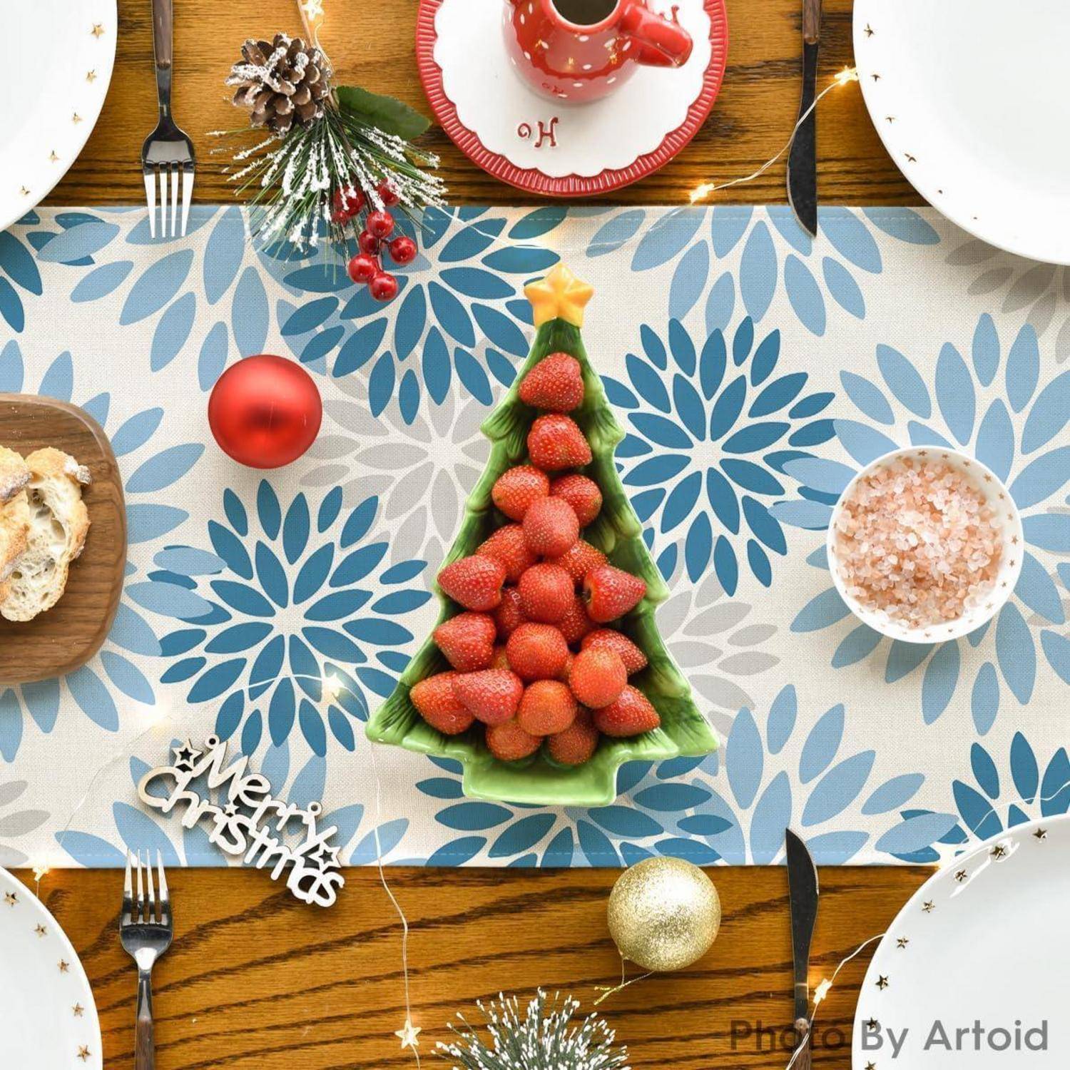 Eine weihnachtlich gestaltete Tischdecke zeigt einen baumförmigen Teller mit Erdbeeren, festliche Dekorationen und eine Tasse mit dem Buchstaben ‚H' auf einem Unterteller.