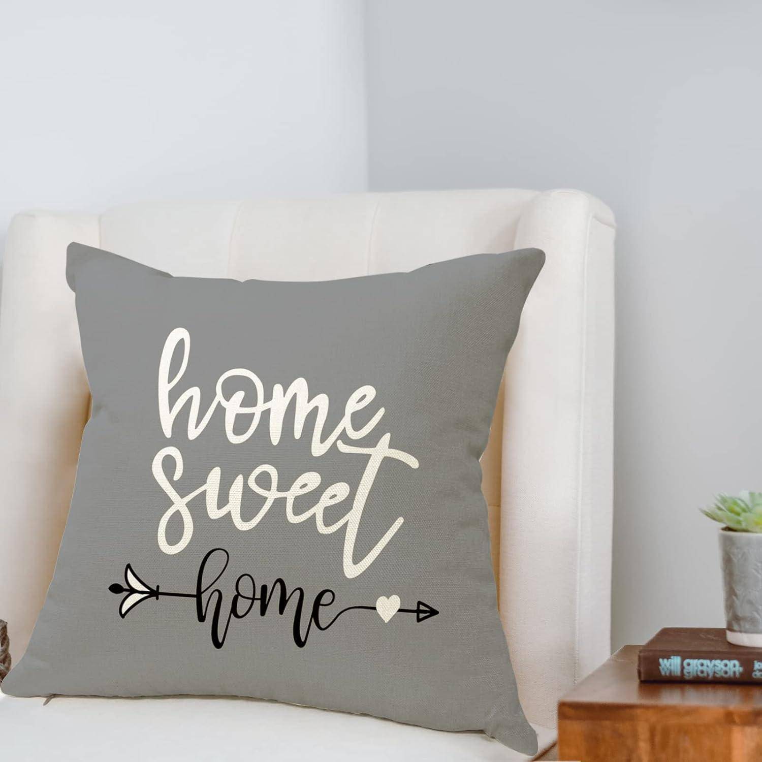 Ein grauer Kissenbezug mit dem Aufdruck 'home sweet home' liegt auf einem beigen Sessel. Eine kleine Pflanze und ein Holztisch befinden sich in der Nähe und schaffen eine gemütliche Atmosphäre.
