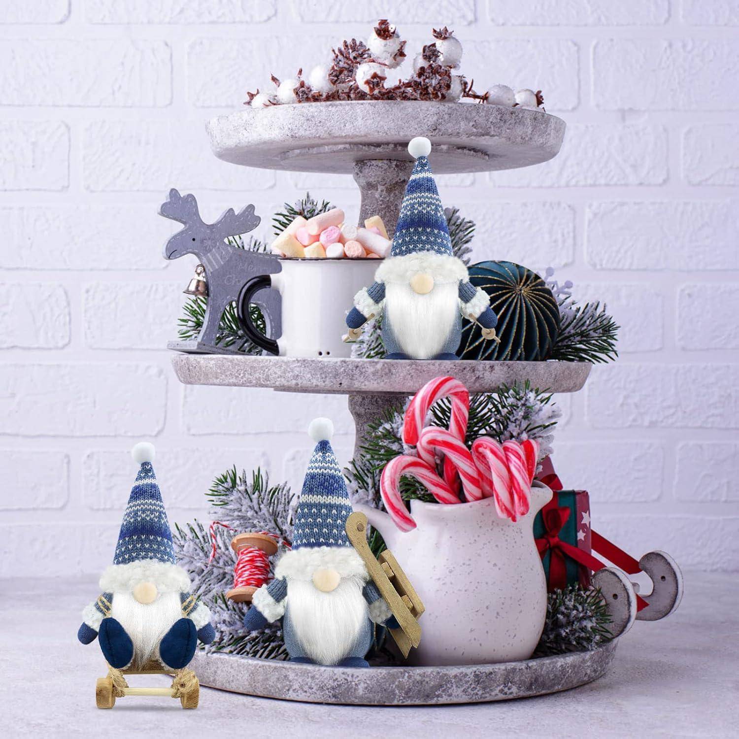 Zweistufiges Tablett mit Weihnachtswichteln, festlichen Tassen, Zuckerstangen und Tannendekorationen vor einem weißen Ziegelwandhintergrund.