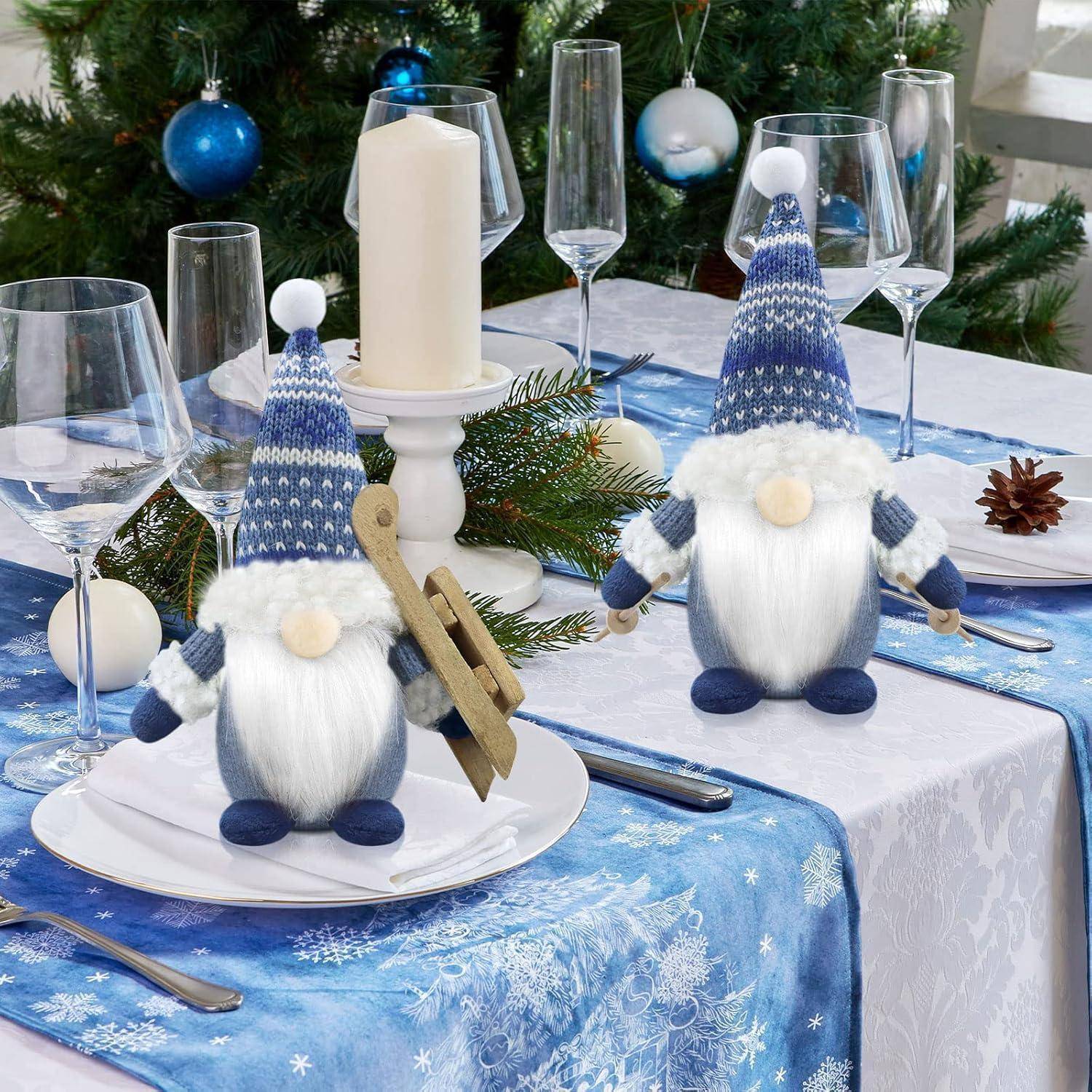 Festliche Tischdecke mit zwei blauen und weißen Gnom-Figuren, umgeben von Kieferndekorationen und Kerzen, vor einem Weihnachtsbaum.