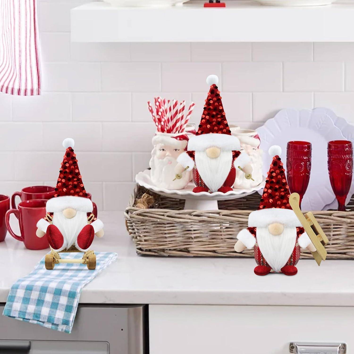 Drei Gnome mit roten Hüten und weißen Bärten sitzen auf der Küchentheke. Sie sind umgeben von festlichen Dekorationen, darunter rote Tassen und weiße Geschirrstücke.