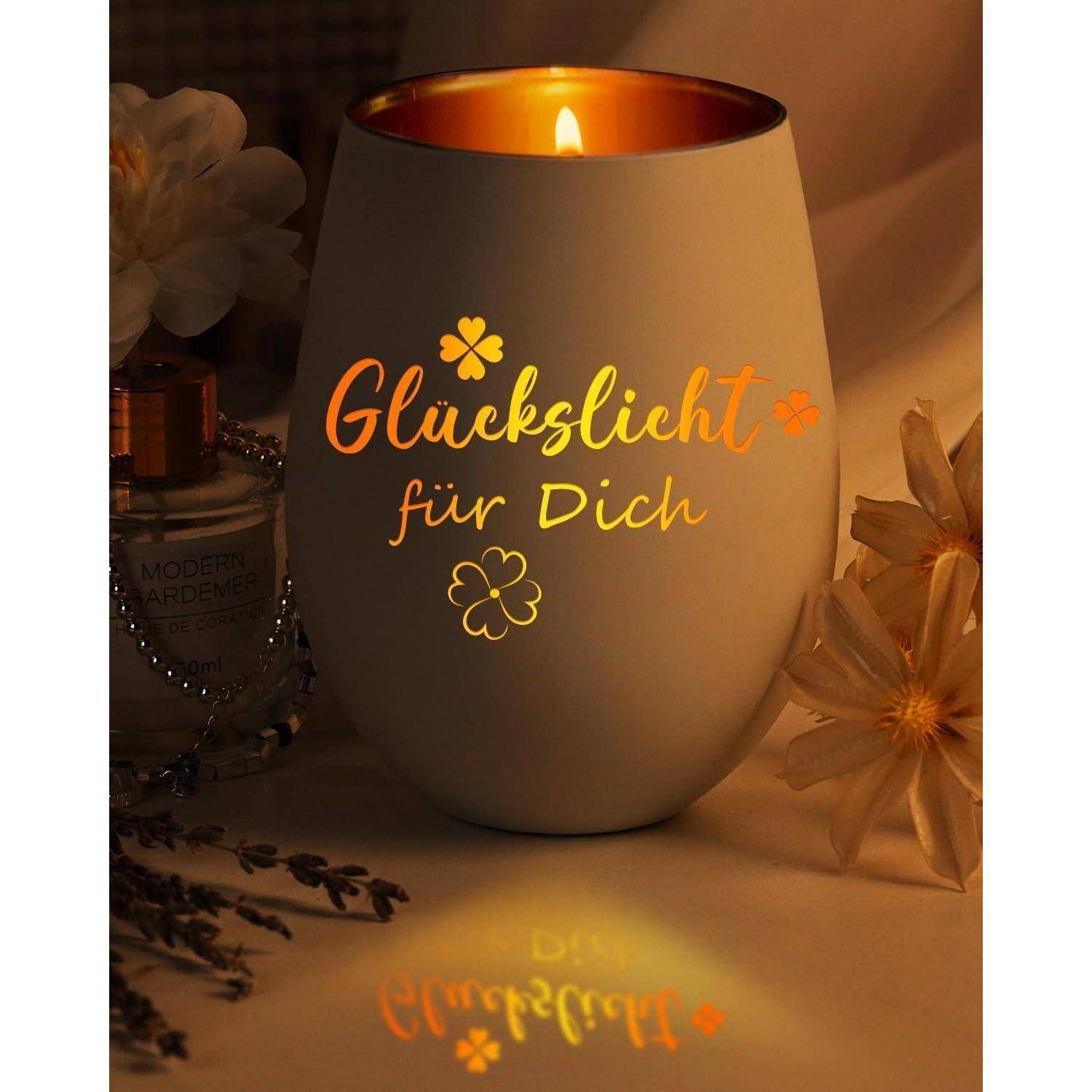 Eine brennende Kerze mit der Aufschrift 'Glückslicht für Dich', umgeben von Blumen und Parfüm, schafft eine warme und gemütliche Atmosphäre.