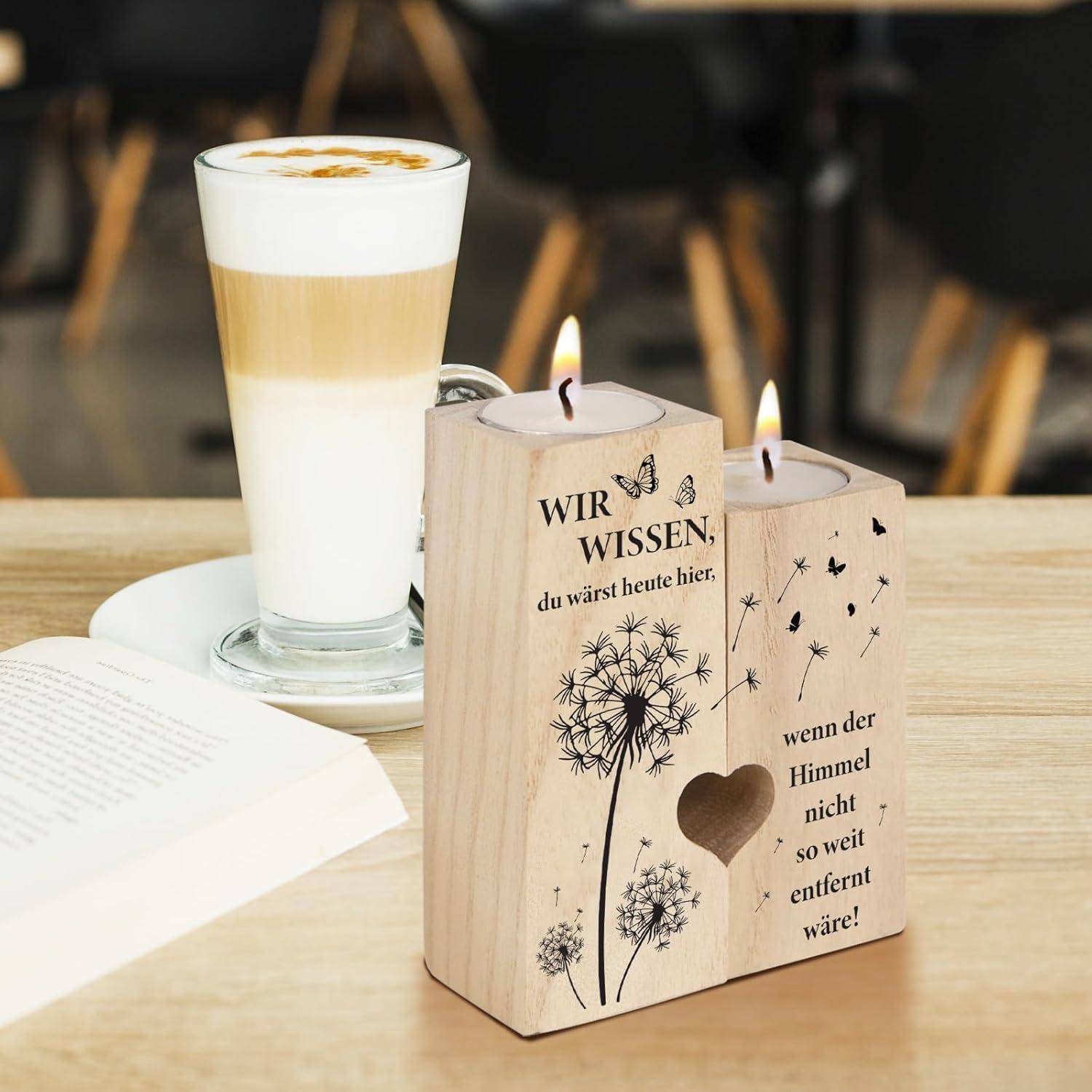 Eine Latte in einer Glastasse auf einem Unterteller neben einem aufgeschlagenen Buch und einem Kerzenhalter aus Holz mit einem Löwenzahnmotiv und deutschem Text.