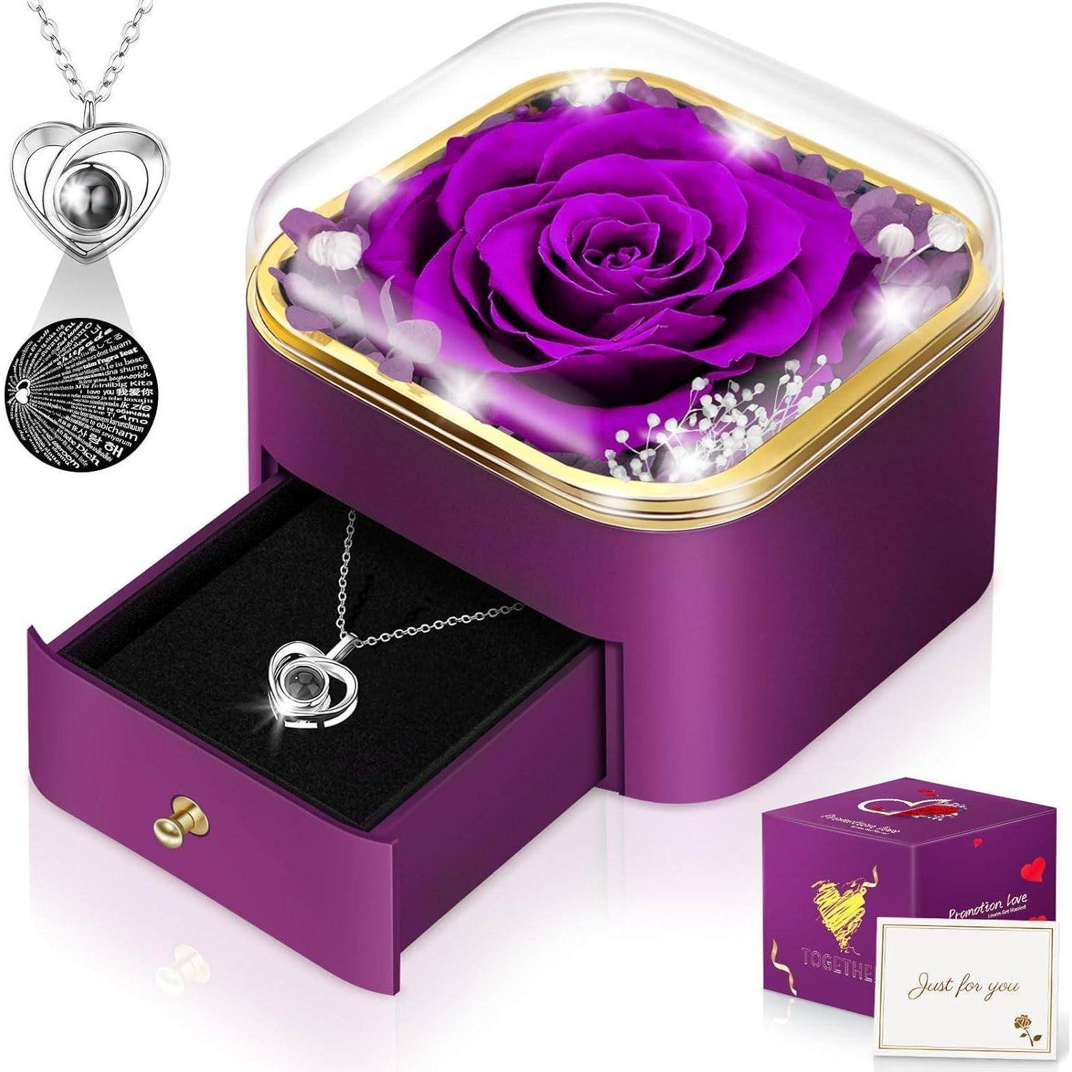 Eine lebendige, konservierte violette Rose in einer Schachtel mit transparentem Deckel, in deren Schublade eine herzförmige Halskette liegt, was ein Geschenkset andeutet.
