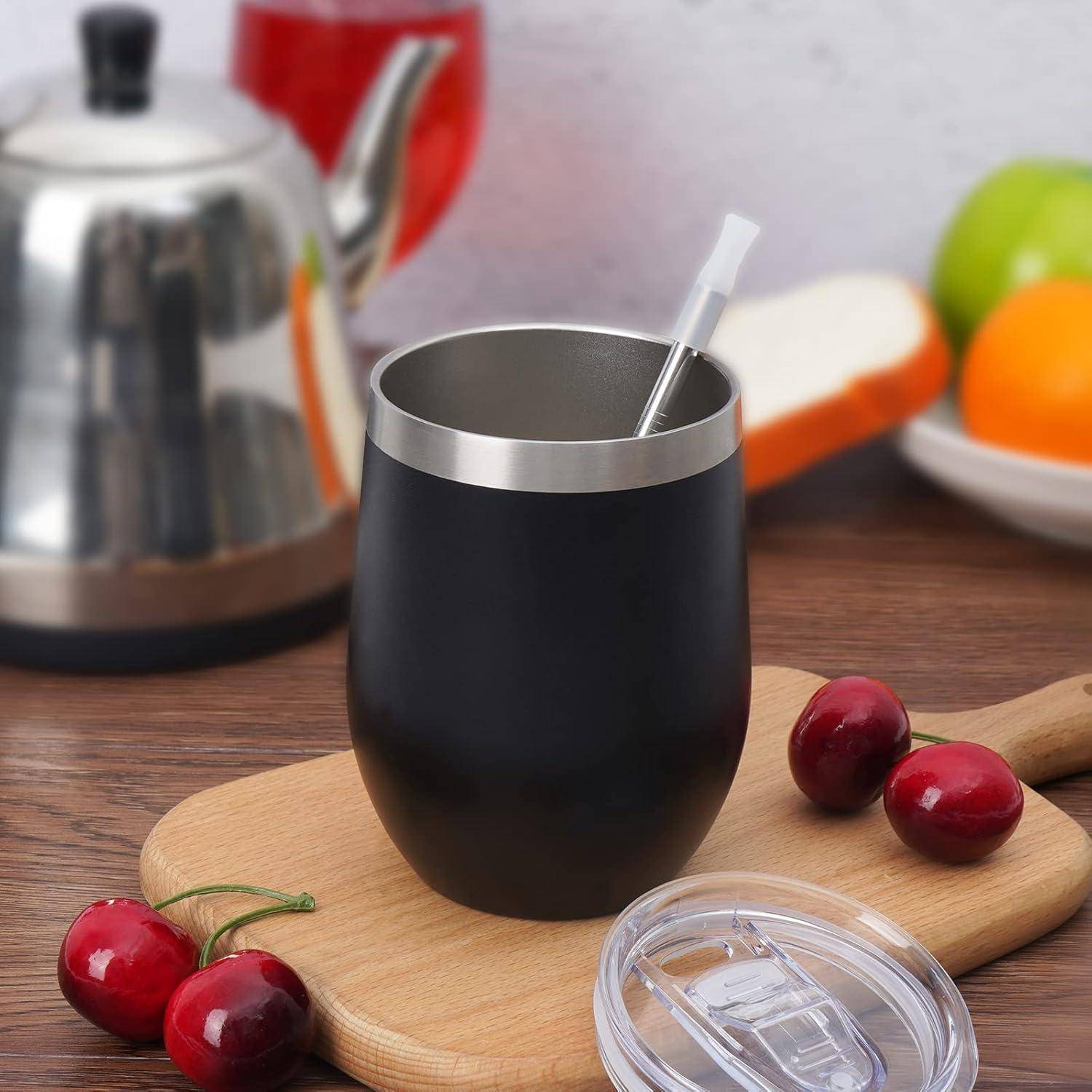 Eine schwarze isolierte Tasse mit Strohhalm und Deckel steht auf einem Holzschneidebrett, umgeben von frischen Kirschen; ein Wasserkocher und Früchte befinden sich im Hintergrund.