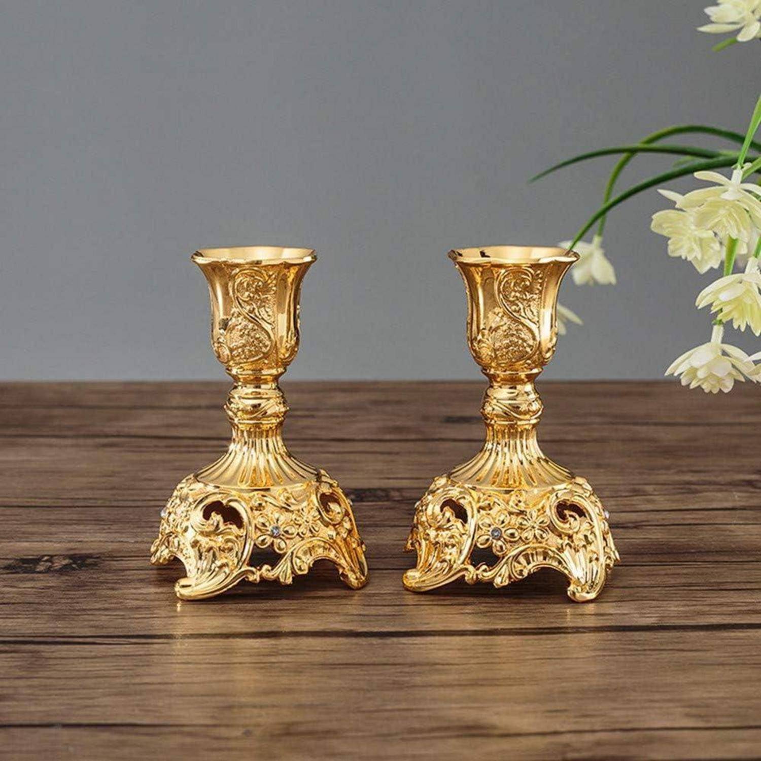 Zwei kunstvoll gearbeitete Kerzenhalter aus Gold mit komplexen Blumenmustern stehen auf einer Holzoberfläche, begleitet von weißen Blumen im Hintergrund.
