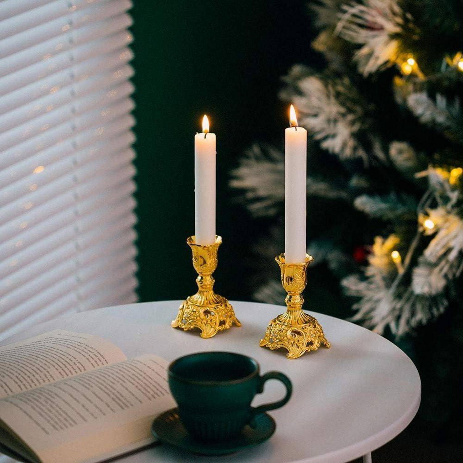 Angezündete Kerzen in kunstvollen Haltern auf einem runden Tisch mit einem aufgeschlagenen Buch und einer grünen Teetasse, in der Nähe eines Fensters und eines Weihnachtsbaums.