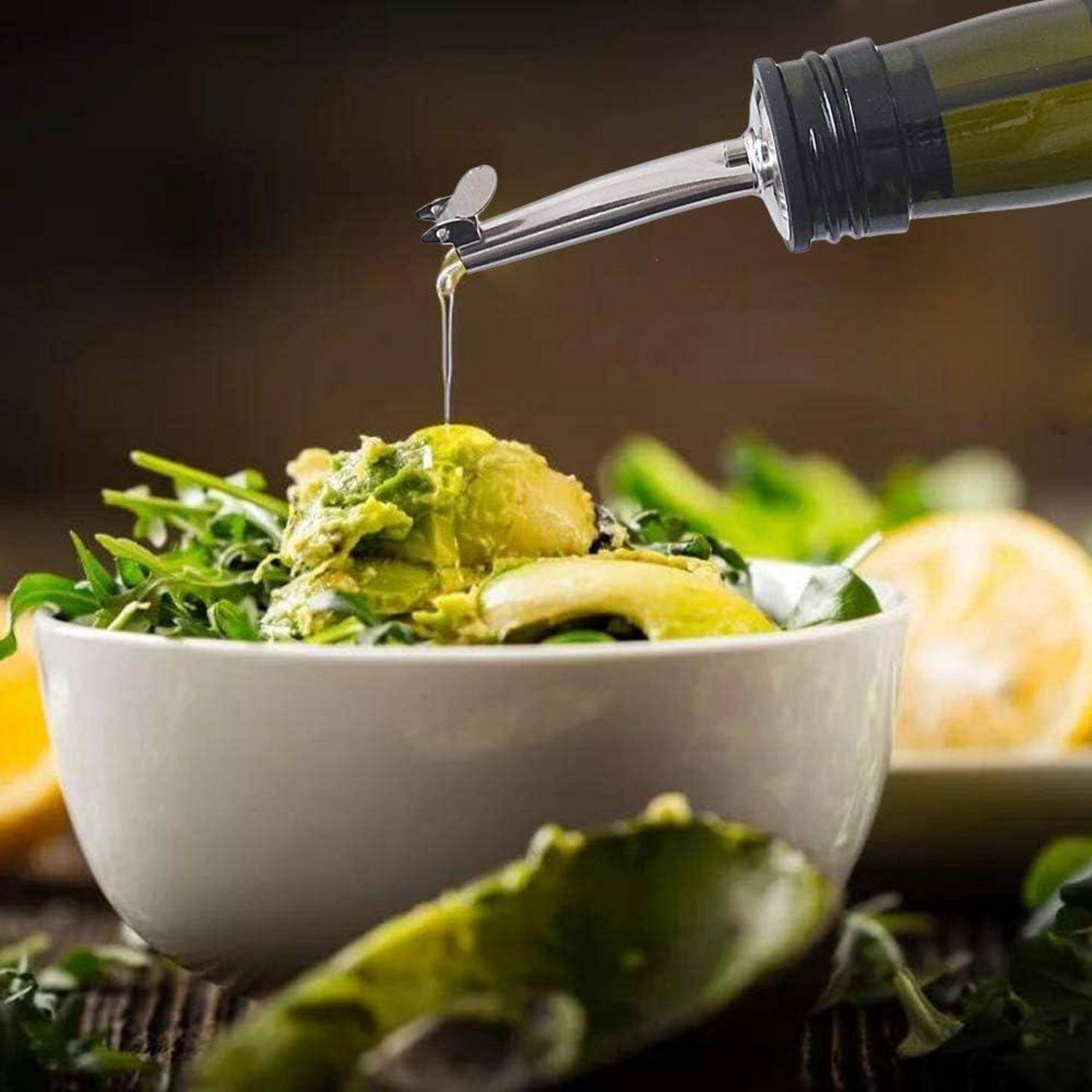 Ein Salat, der mit Olivenöl aus einer Flasche beträufelt wird. Die Schüssel enthält gemischte Blattсалаты, Avocadoscheiben und Zitronenspalten vor einem unscharfen Hintergrund.