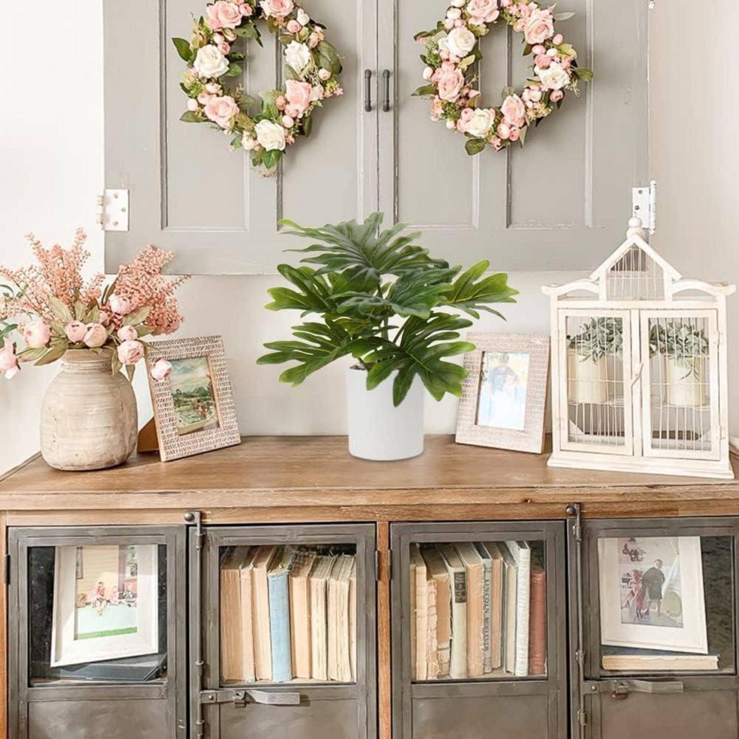 Eine gemütliche Wohnzimmereinrichtung mit einem Holzschrank, Büchern, gerahmten Fotos und Vasen mit rosa Blumen. Zwei Blumenkränze hängen an der Wand.
