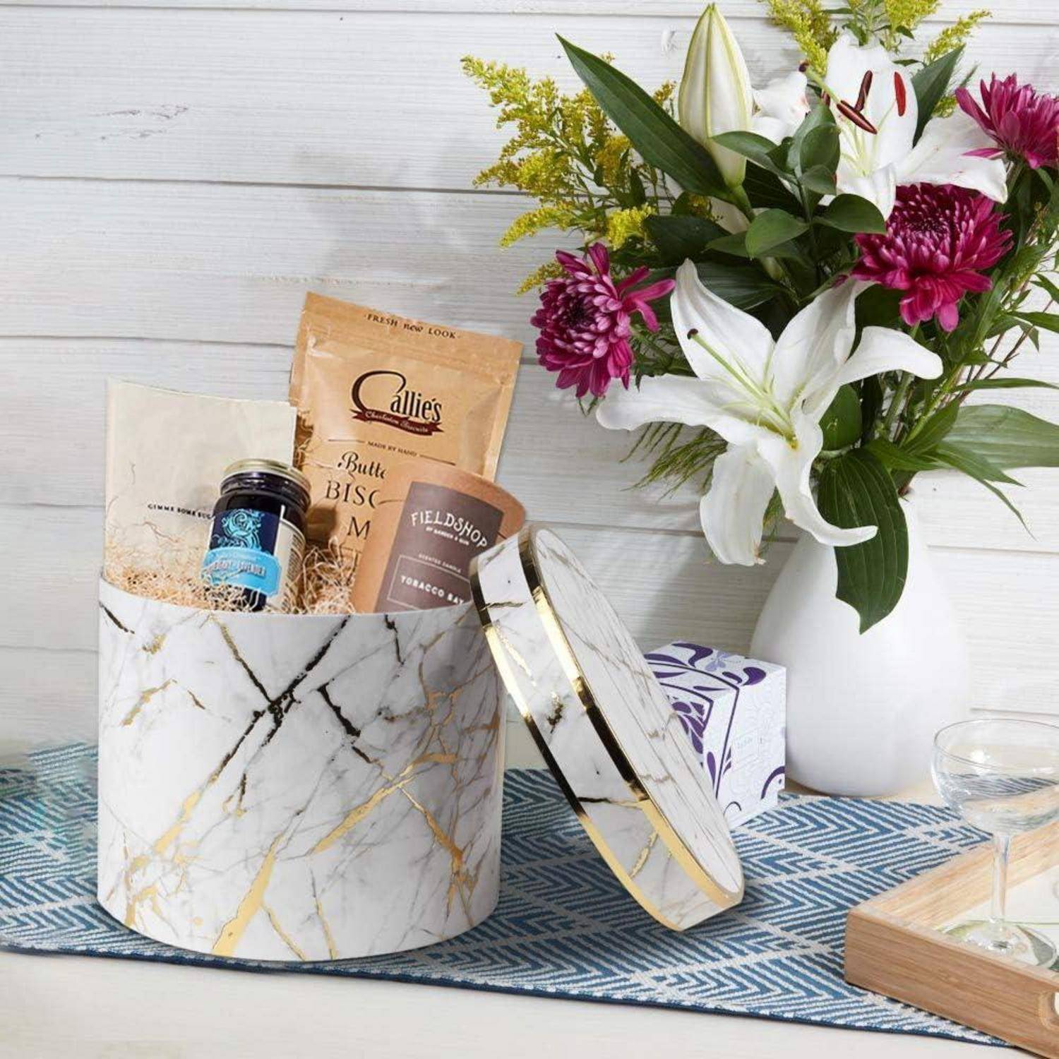 Eine dekorative Marmorbox, gefüllt mit Gourmet-Snacks, ist auf einem Tisch neben einer Vase mit weißen Lilien und lila Blumen geöffnet und schafft eine elegante Geschenkpräsentation.