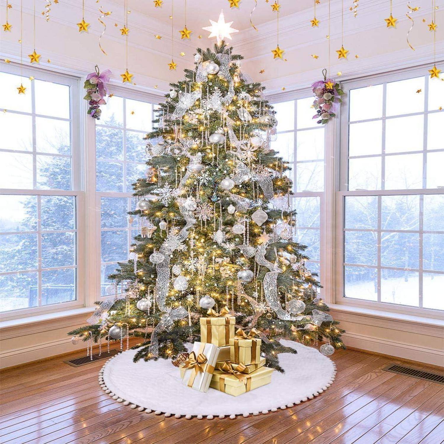 Ein großer, geschmückter Weihnachtsbaum mit Ornamenten und Lichtern steht in einem hellen Raum, umgeben von Fenstern, die eine verschneite Landschaft zeigen.