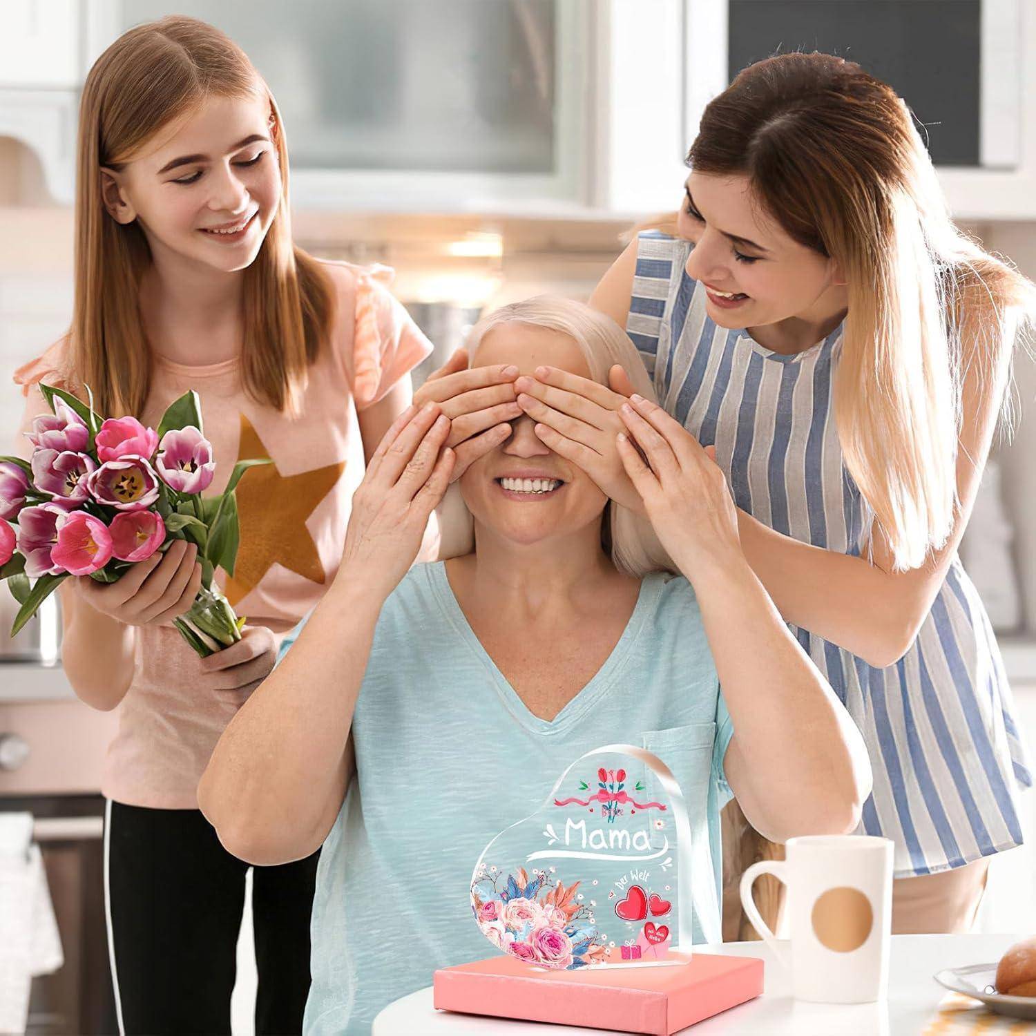 Eine lächelnde Frau sitzt an einem Küchentisch, ihre Augen werden von einer jüngeren Frau verdeckt. Eine weitere junge Frau steht in der Nähe und hält einen Blumenstrauß.