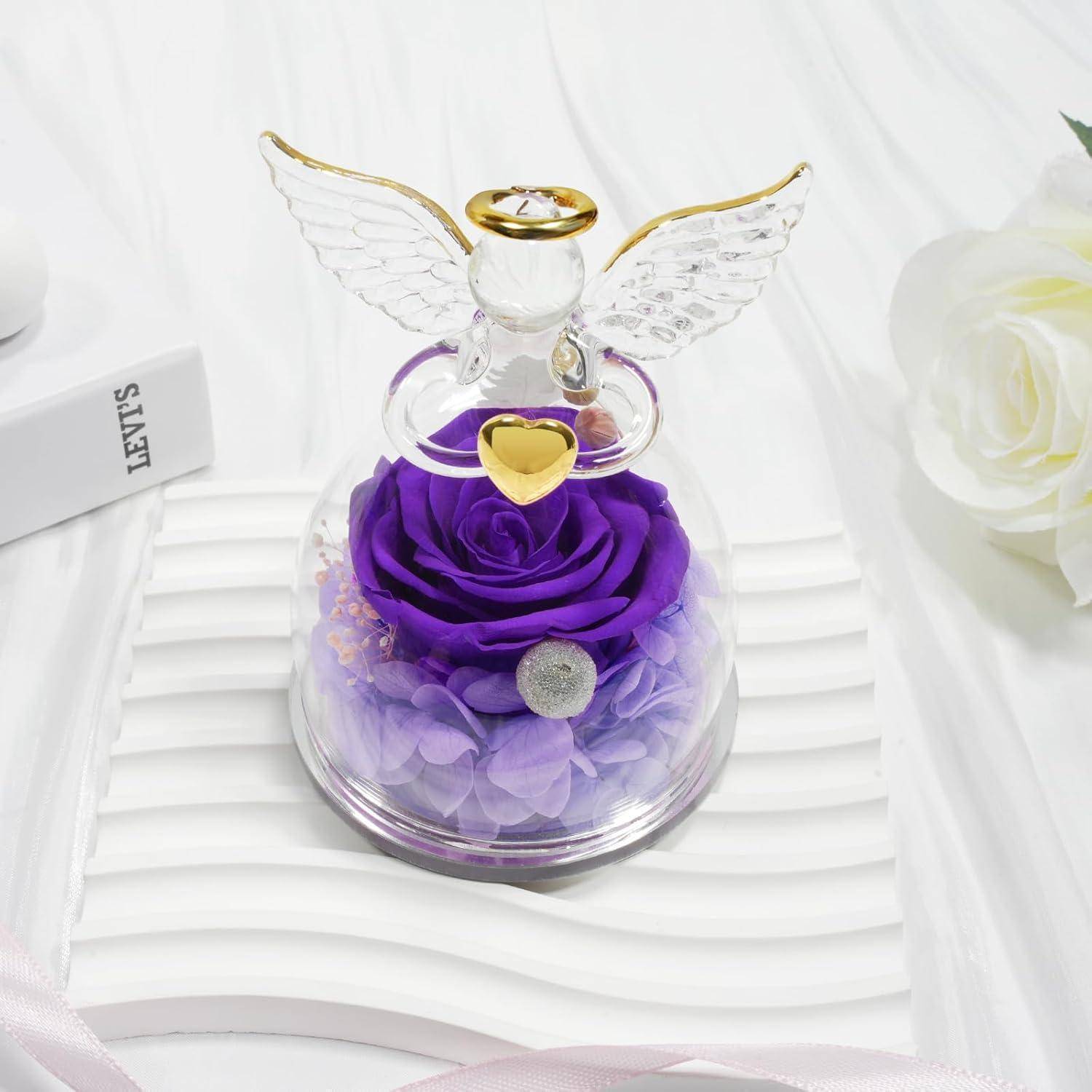 Eine Glasengelfigur mit goldenen Akzenten sitzt über einer konservierten lila Rose und Hortensien in einer durchsichtigen Glaskuppel auf einer gestreiften Oberfläche.