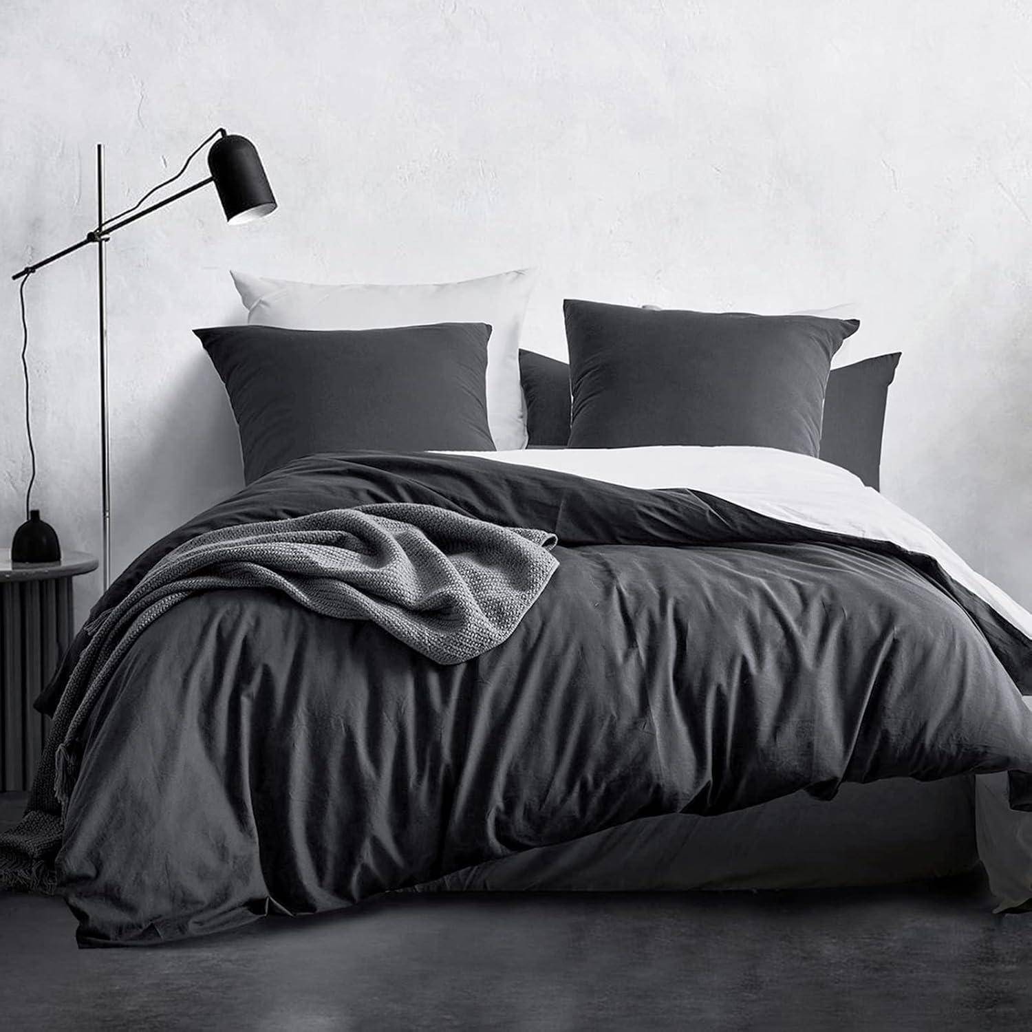 Ein ordentlich aufgemachtes Bett mit einer schwarzen Stehlampe daneben. Das Bett ist mit dunkelgrauer Bettwäsche und Kissen ausgestattet, und eine strukturierte graue Decke ist darüber drapiert. Die Umgebung vermittelt einen modernen, minimalistischen Stil.