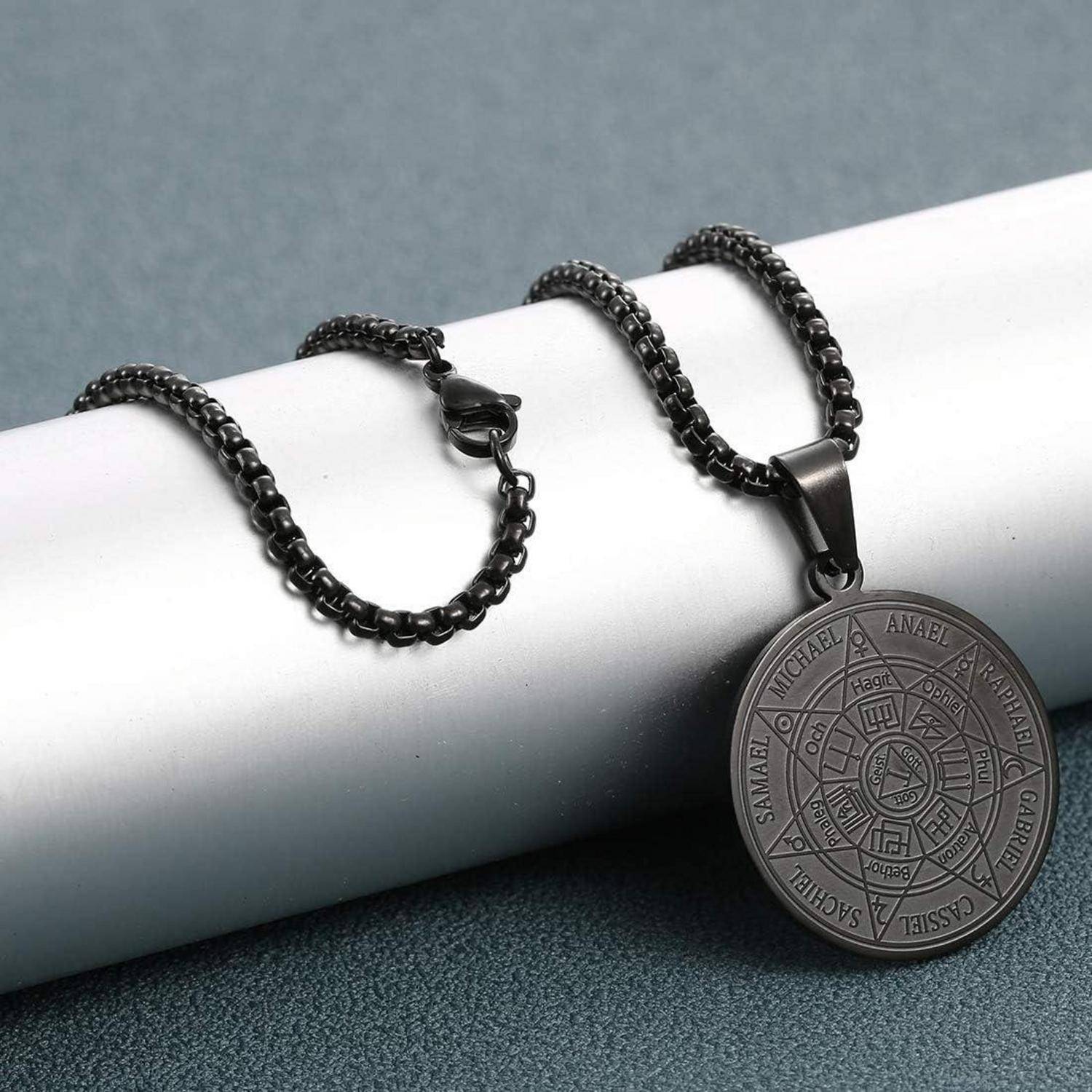 Eine schwarze Metallkette mit einem runden Anhänger, der Engelnamen und -symbole trägt, ausgelegt auf einer strukturierten grauen Oberfläche.