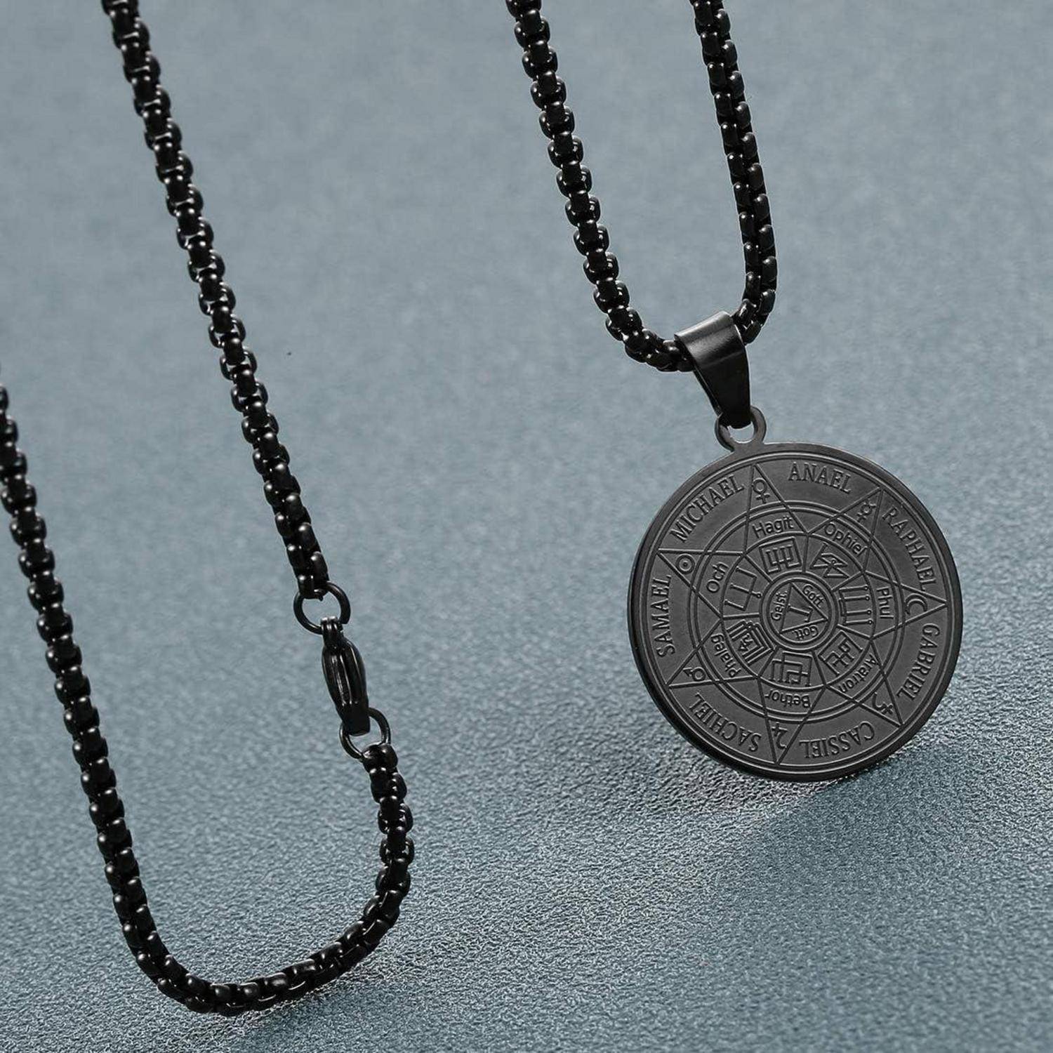 Eine schwarze Anhängerkette mit einem kreisförmigen Medaillon, das detaillierte Gravuren von Namen und Symbolen trägt, präsentiert auf einer strukturierten Oberfläche.