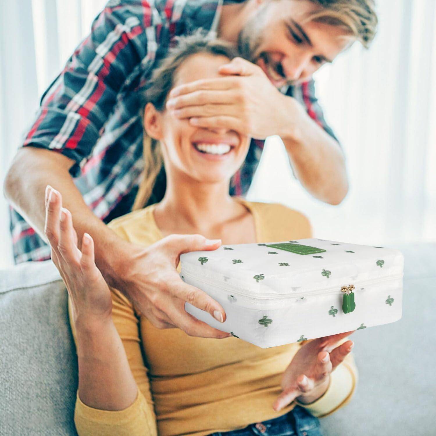 Ein Mann überrascht eine Frau, indem er ihre Augen von hinten bedeckt, während sie nach einer weißen Geschenkbox mit grünen Kleeblättern greift, die auf einem beigen Sofa liegt.