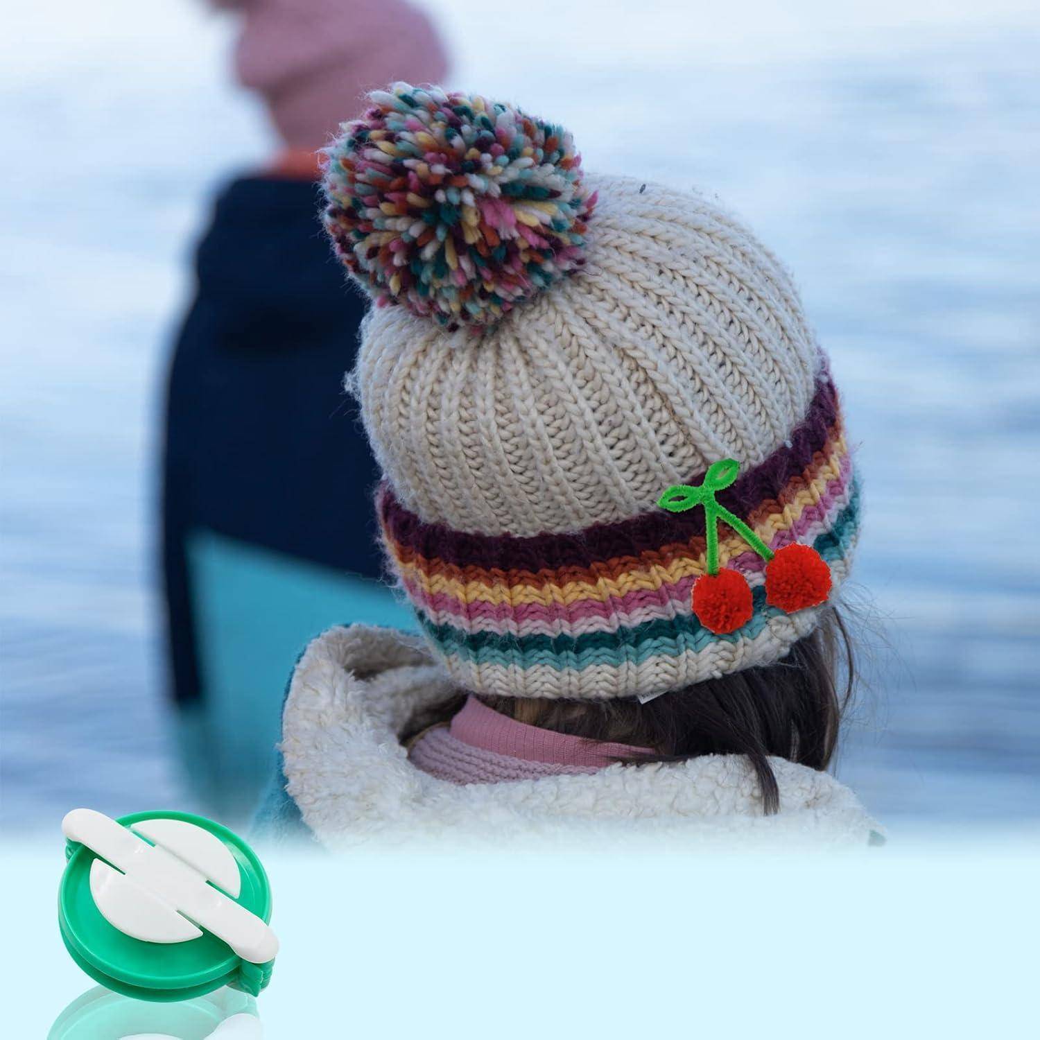 Ein Kind mit einer bunten gestrickten Mütze mit Kirschverzierung steht an ruhigem Wasser, während ein Erwachsener im Hintergrund zu sehen ist. Ein grüner Gegenstand befindet sich im Vordergrund.